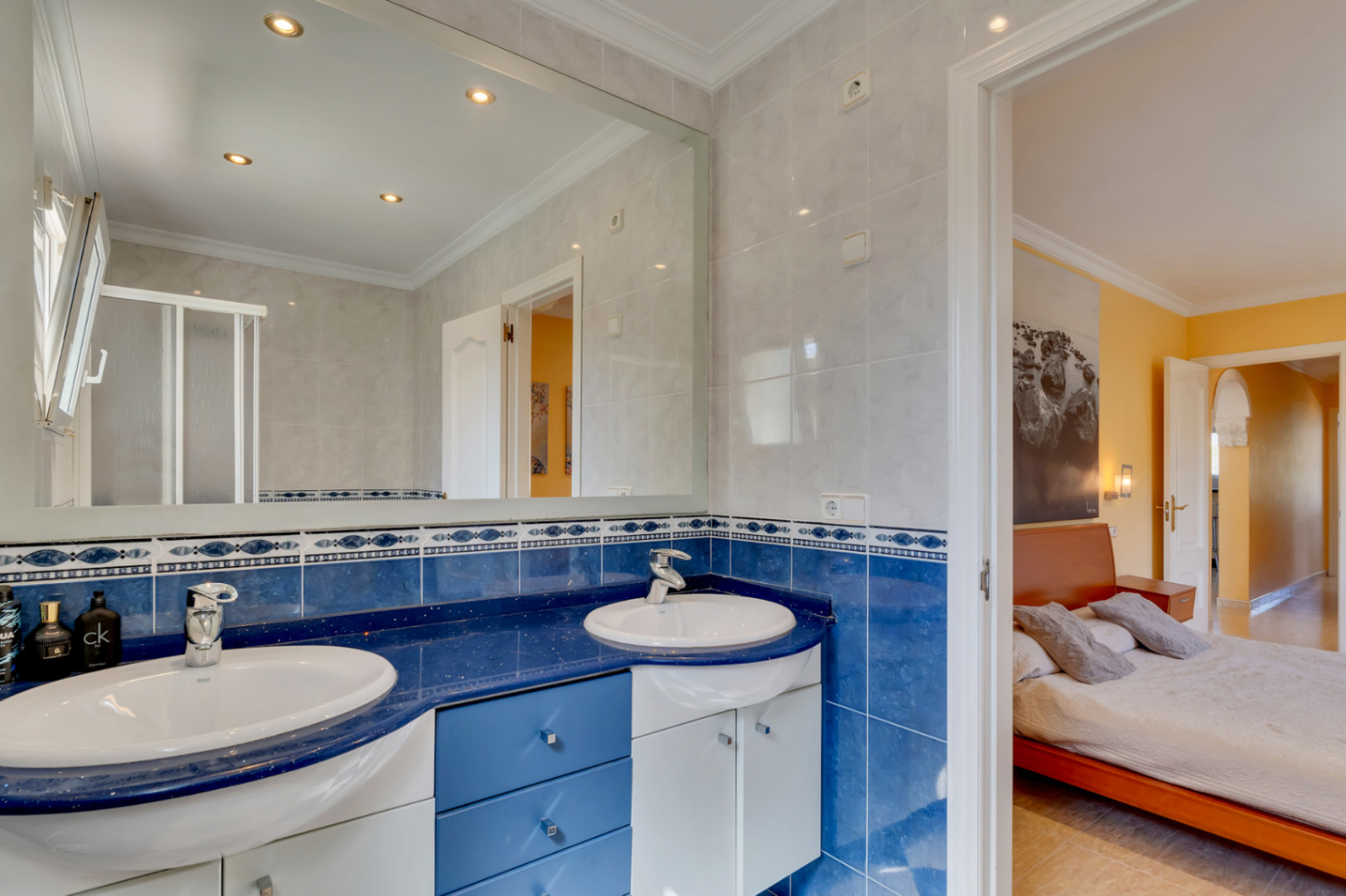 Badezimmer mit Doppelwaschtisch auf blauer Arbeitsplatte, dekorativen Fliesen und großem Spiegel. Direkter Zugang zum Schlafzimmer mit gelben Wänden.