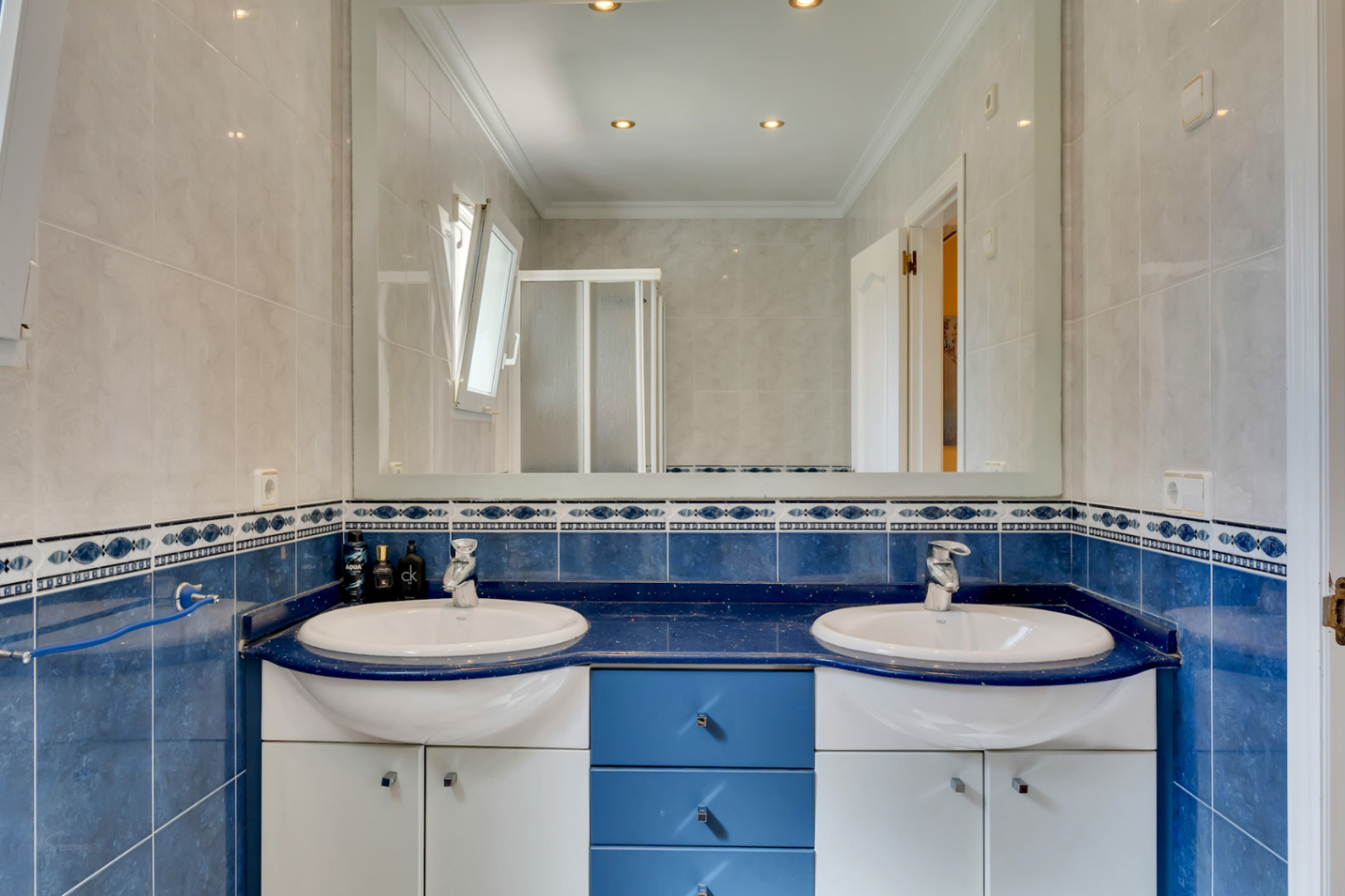 Badezimmer mit Doppelwaschtisch auf blauer Arbeitsplatte, verchromten Armaturen und viel Stauraum. Blaue und weiße Wandfliesen ergänzen den großflächigen Spiegel und die Spotbeleuchtung.
