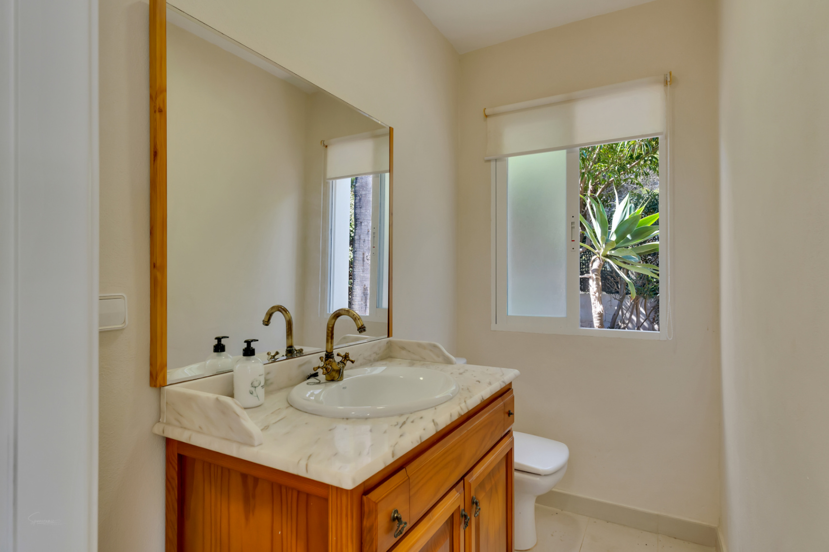 Badezimmer mit Holzwaschtisch, Marmorplatte und Armaturen im klassischen Stil. Ein Fenster sorgt für natürliches Licht und Ausblick auf die Gartenpflanzen.