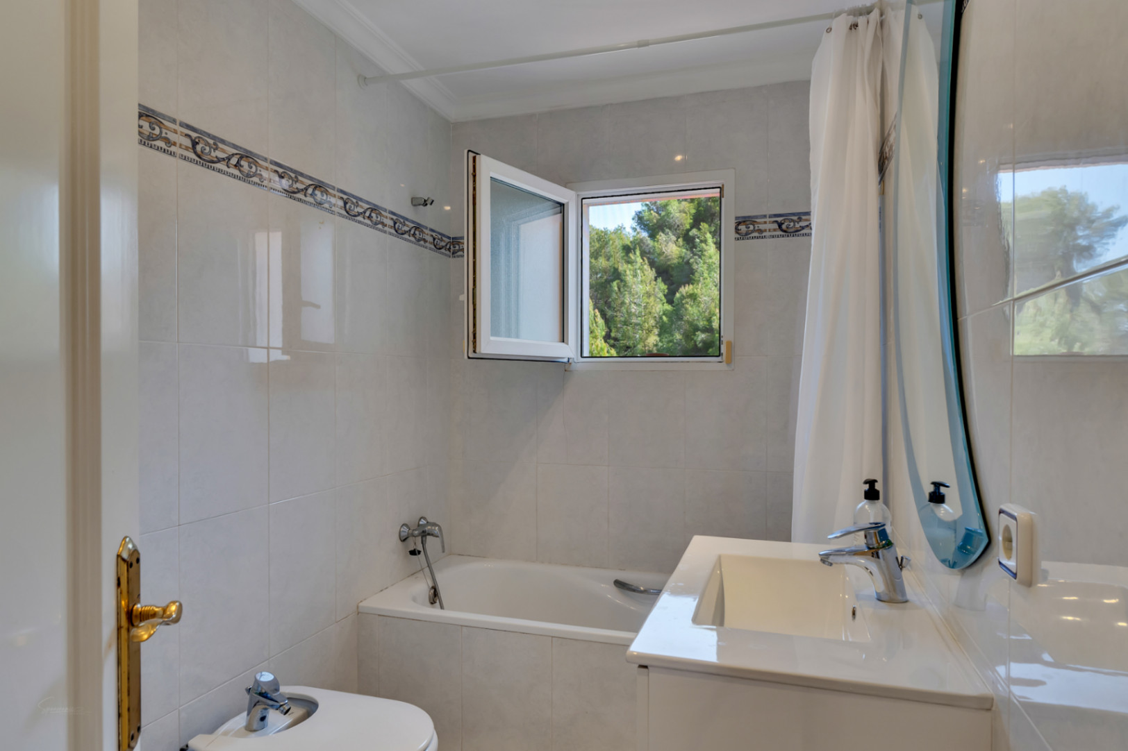 Funktionales Badezimmer mit Einbaubadewanne, Bidet und weißem Waschtisch. Ein Fenster öffnet sich zum Blick ins Grüne, ergänzt durch helle Fliesen mit Zierbordüre.