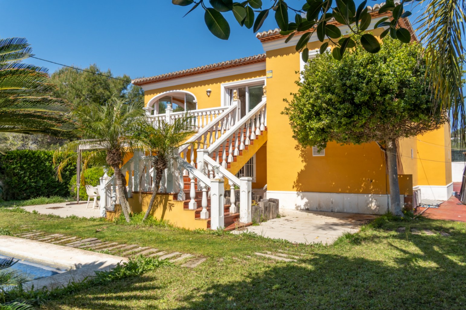 Gelbe Villenfassade mit einer Außentreppe und weißer Balustrade, die zur oberen Terrasse führt. Mediterraner Garten mit Palmen, Rasenfläche und Poolbereich.