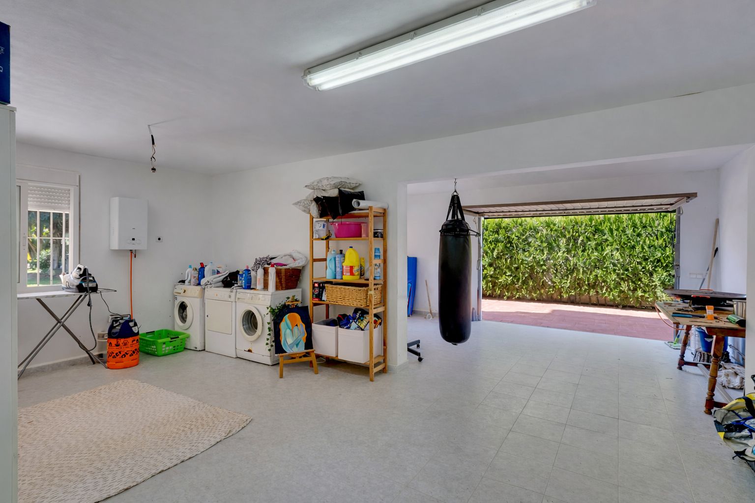 Großzügiger Mehrzweckraum mit Waschküche, Lagerregalen und Fitnessbereich inklusive Boxsack, mit direktem Zugang zur Garageneinfahrt und zum Garten.