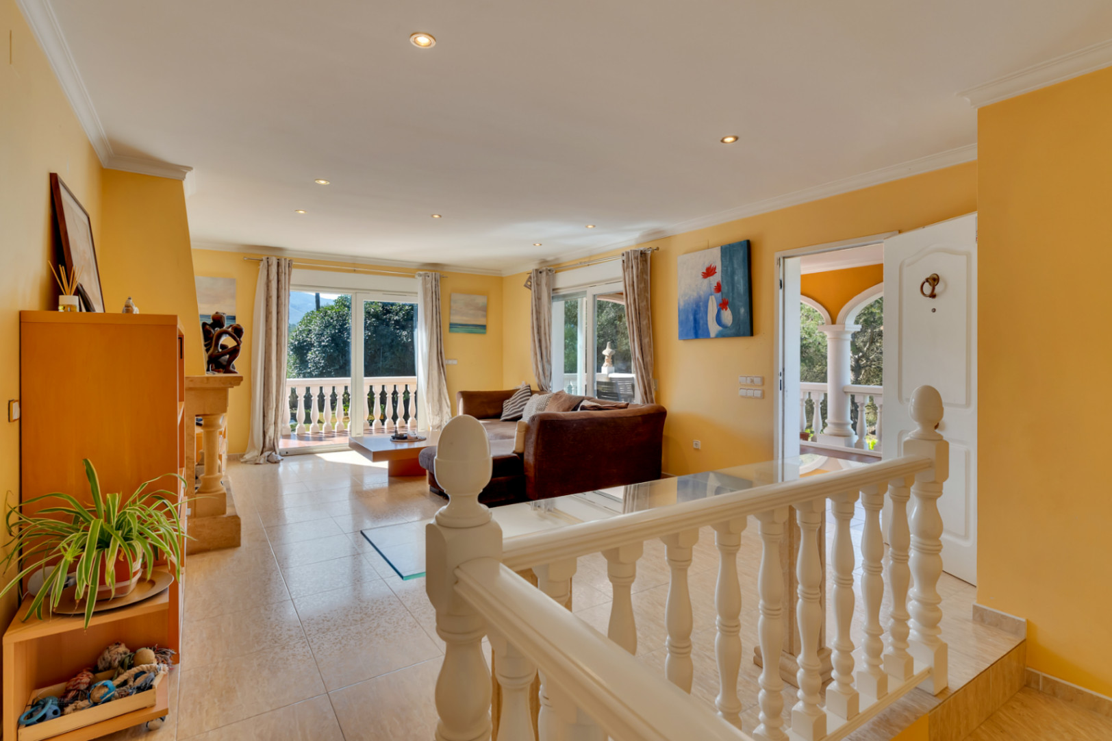 Heller Wohnbereich mit Fliesenboden, Deckeneinbaustrahlern und großen Glasschiebetüren, die auf eine Terrasse mit klassischer Balustrade und Blick ins Grüne führen.