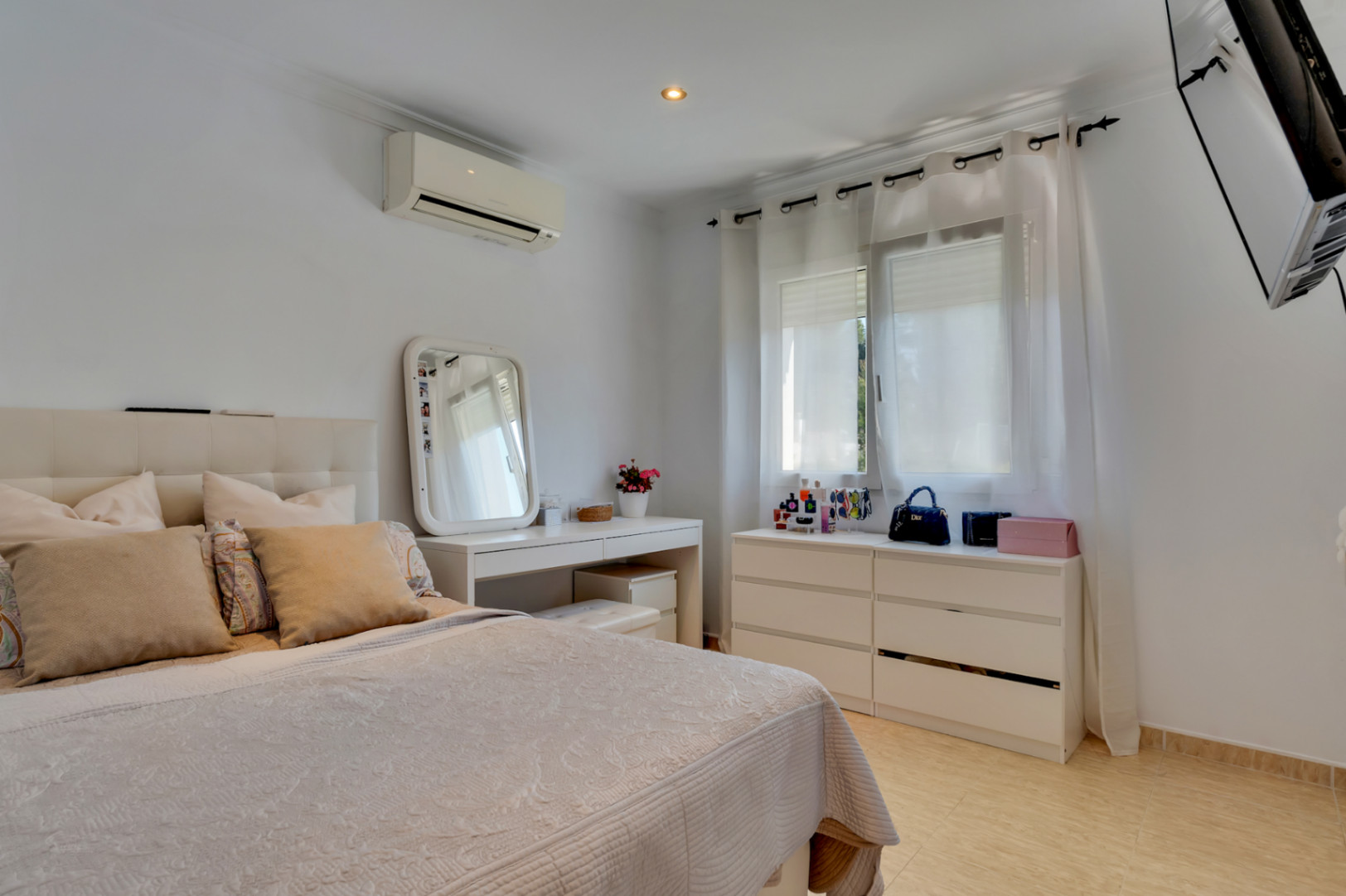 Helles Schlafzimmer mit gepolstertem Kopfteil, weißem Schminktisch und passender Kommode. Ausgestattet mit Klimaanlage, Fliesenboden und wandmontiertem Fernseher.
