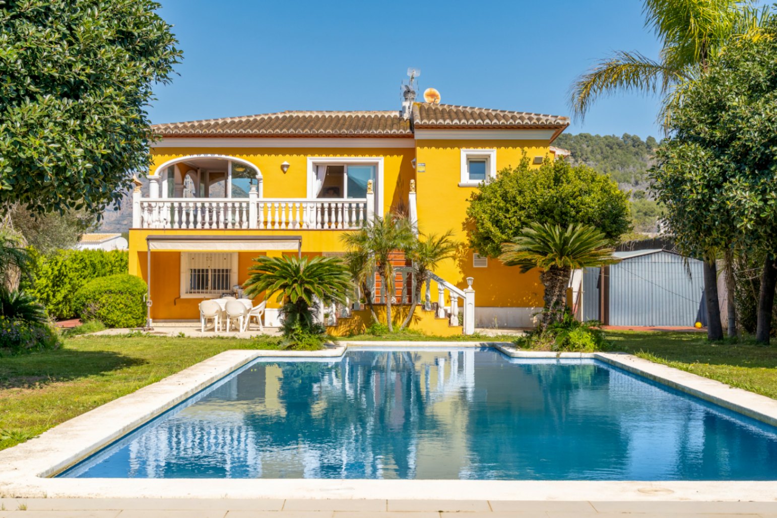 Mediterrane Villa mit gelber Fassade, großem Pool, Terrasse mit weißer Balustrade und einem Garten mit Palmen und weitläufiger Rasenfläche.