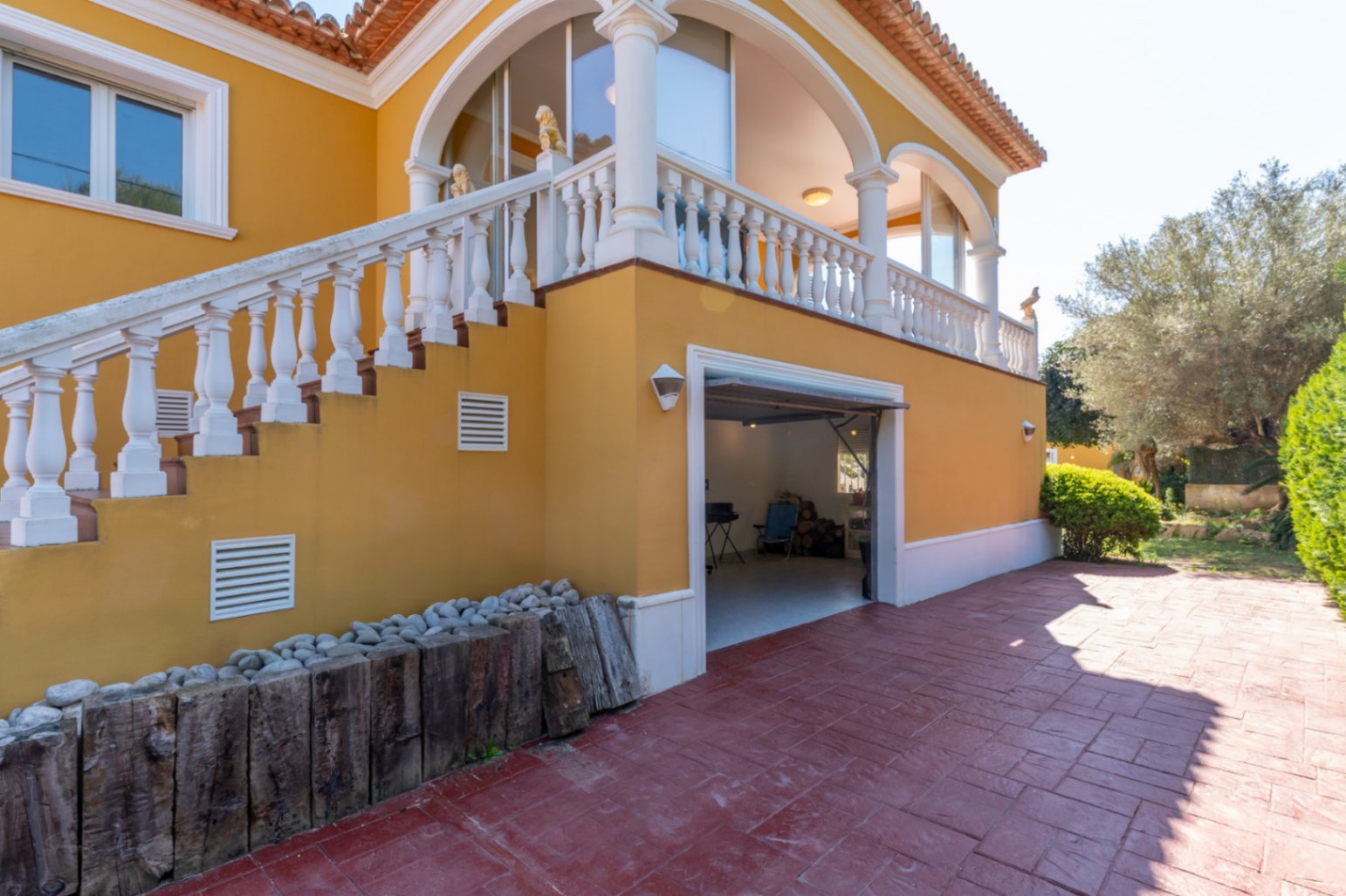Ockerfarbene Fassade mit Außentreppe und weißer Balustrade, die zu einer Bogenterrasse führt. Integrierte Garage und gepflasterter Zufahrtsbereich.