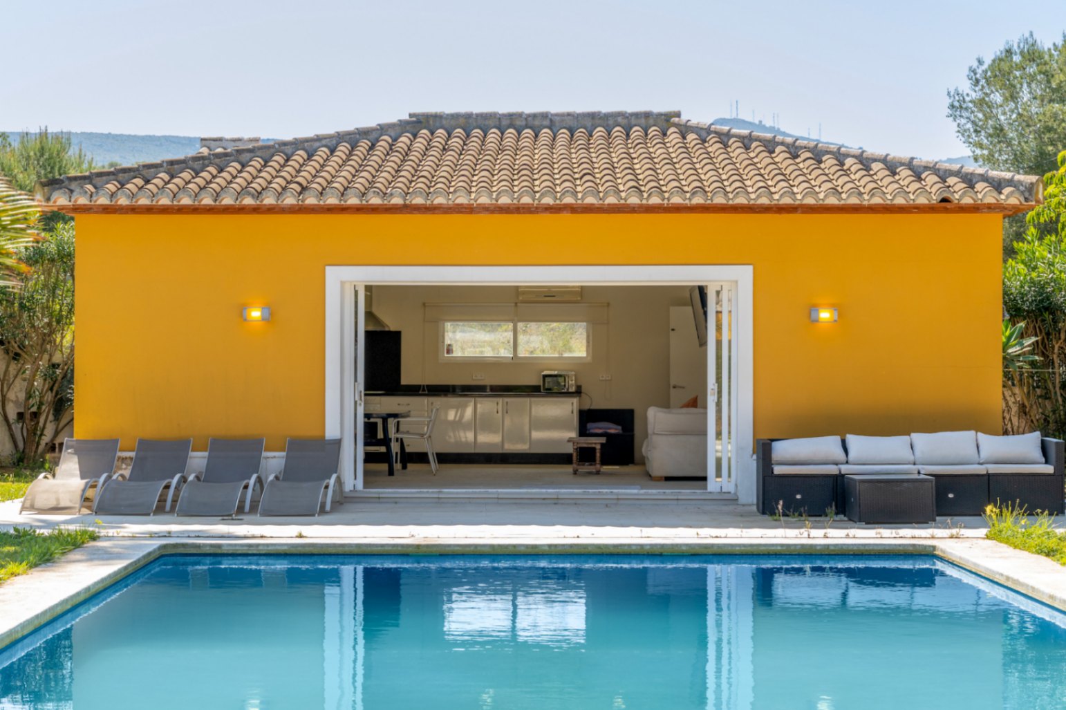 Poolhaus mit ausgestatteter Küche, Wohnbereich und großen Glasschiebetüren, die direkt auf die Sonnenterrasse mit Liegestühlen und Lounge-Möbeln führen.