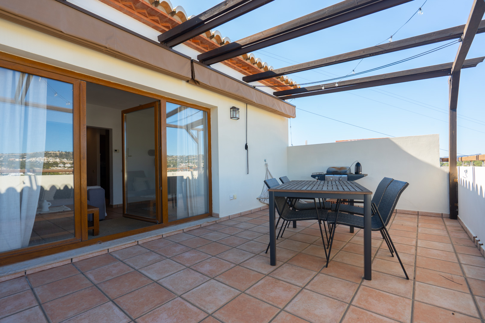 Außenterrasse mit Terrakottafliesen, Essbereich unter einer Holzpergola und Grillstation. Große Glasschiebetüren mit Holzrahmen verbinden den Außenbereich mit dem Wohnraum.