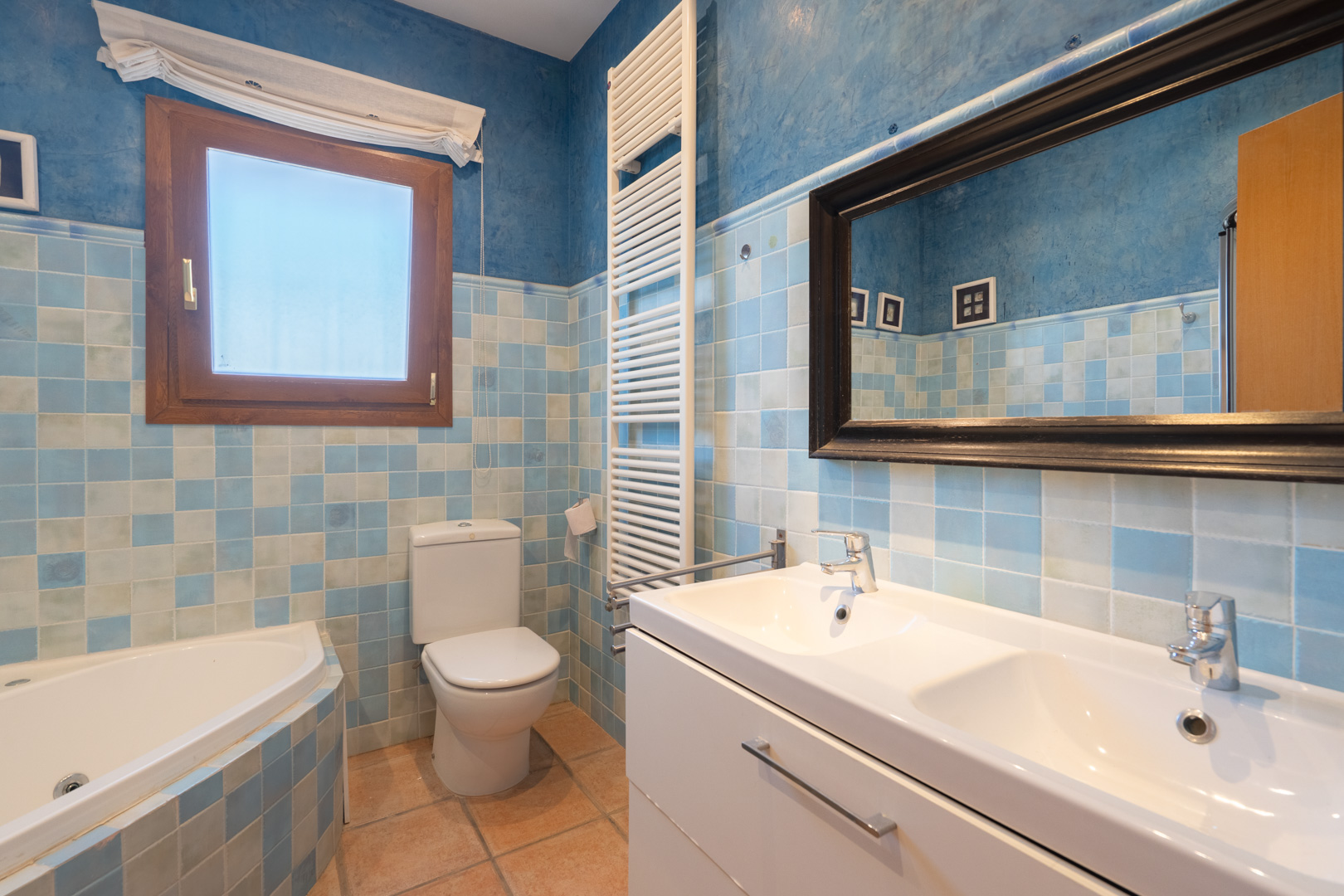 Badezimmer mit blau-weiß karierten Fliesen, Doppelwaschtisch, Eckbadewanne und weißem Handtuchheizkörper vor dekorativ blau gestalteten Wänden.