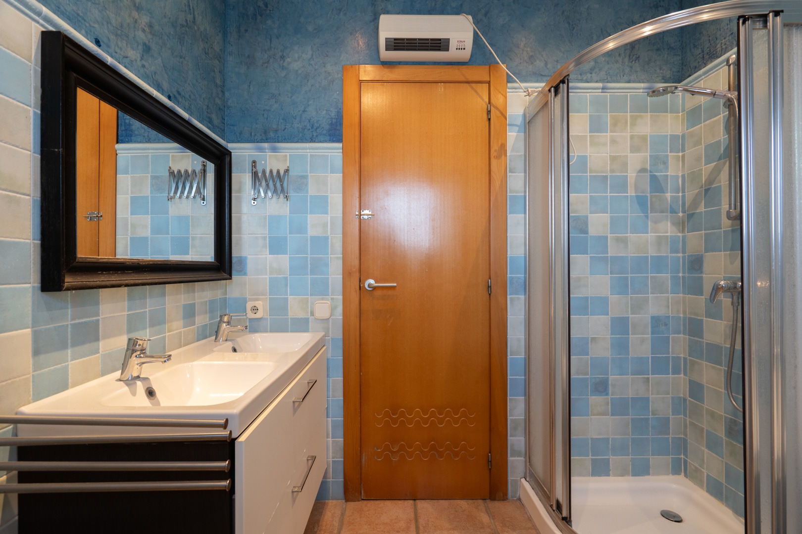 Badezimmer mit Doppelwaschtisch, Eckdusche mit Glasabtrennung und blauem Fliesenspiegel, kombiniert mit einer markanten Holztür und Heizlüfter.