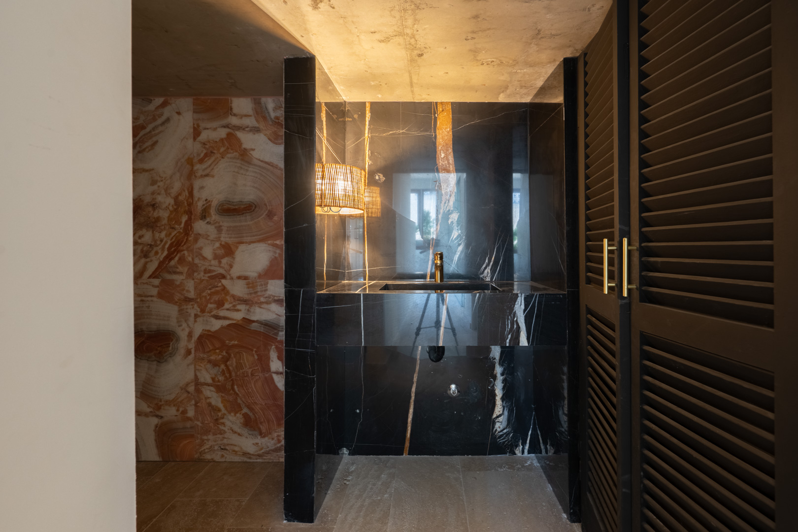 Designer-Gäste-WC mit integriertem Waschbecken aus schwarzem Marmor, goldenen Armaturen und Natursteinwänden. Mit stimmungsvoller Beleuchtung und dunklen Lamellentüren.