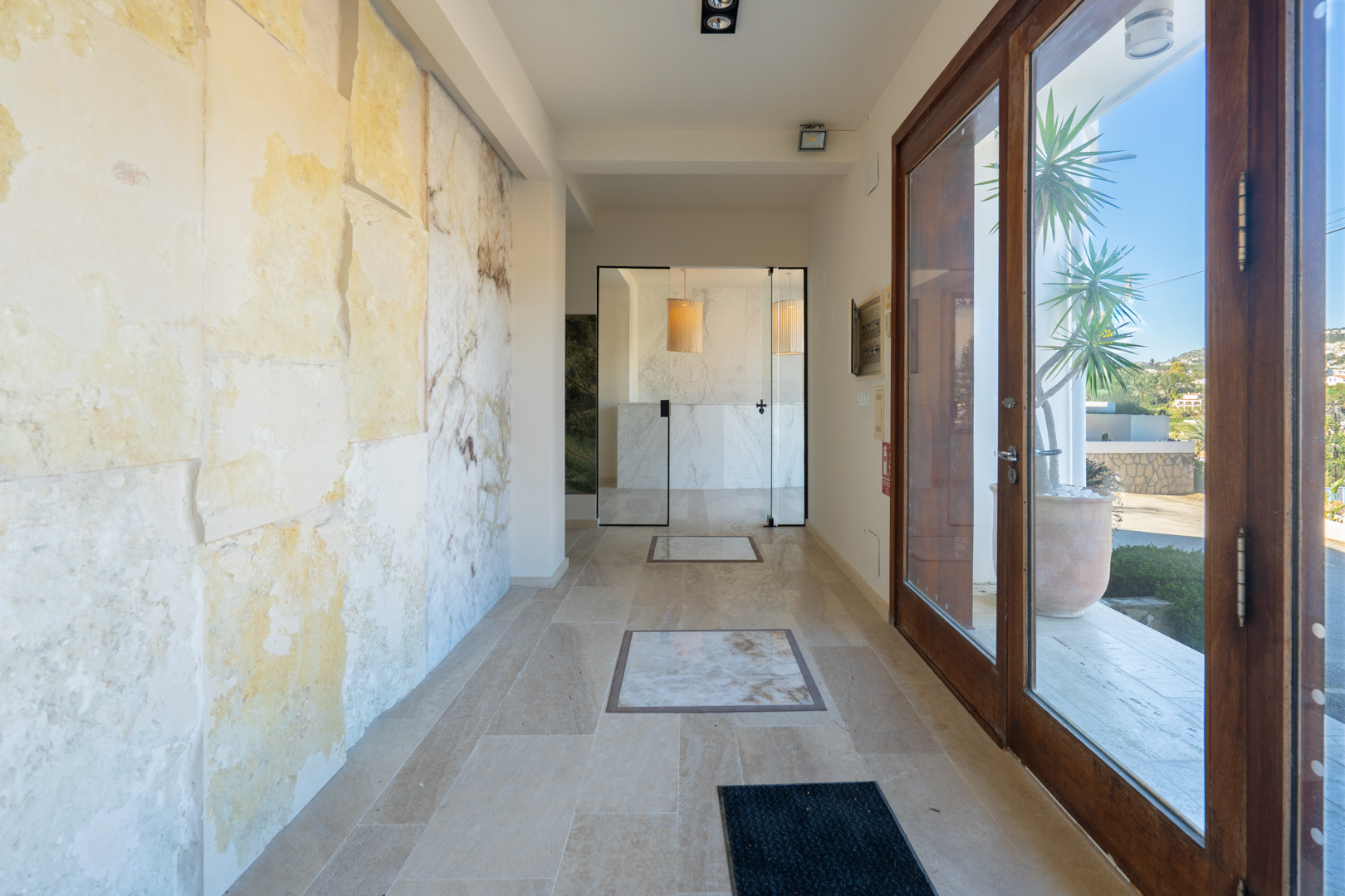 Eingangshalle mit strukturierter Natursteinwand, Fliesenboden mit Marmoreinlagen und großen Glas-Doppeltüren mit Holzrahmen.