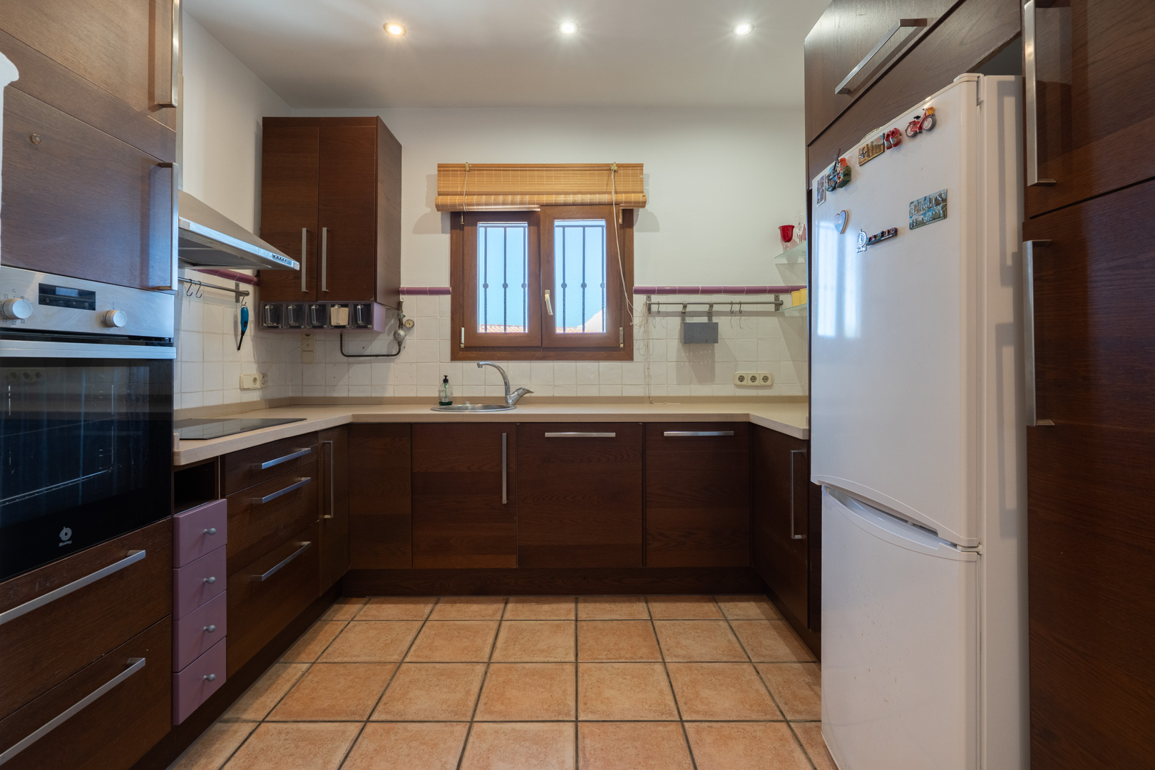 Funktionale Küche mit dunklen Holzschränken, beiger Arbeitsplatte und Terrakotta-Fliesen. Ausgestattet mit Einbaubackofen, weißem Kühlschrank und Deckenstrahlern.
