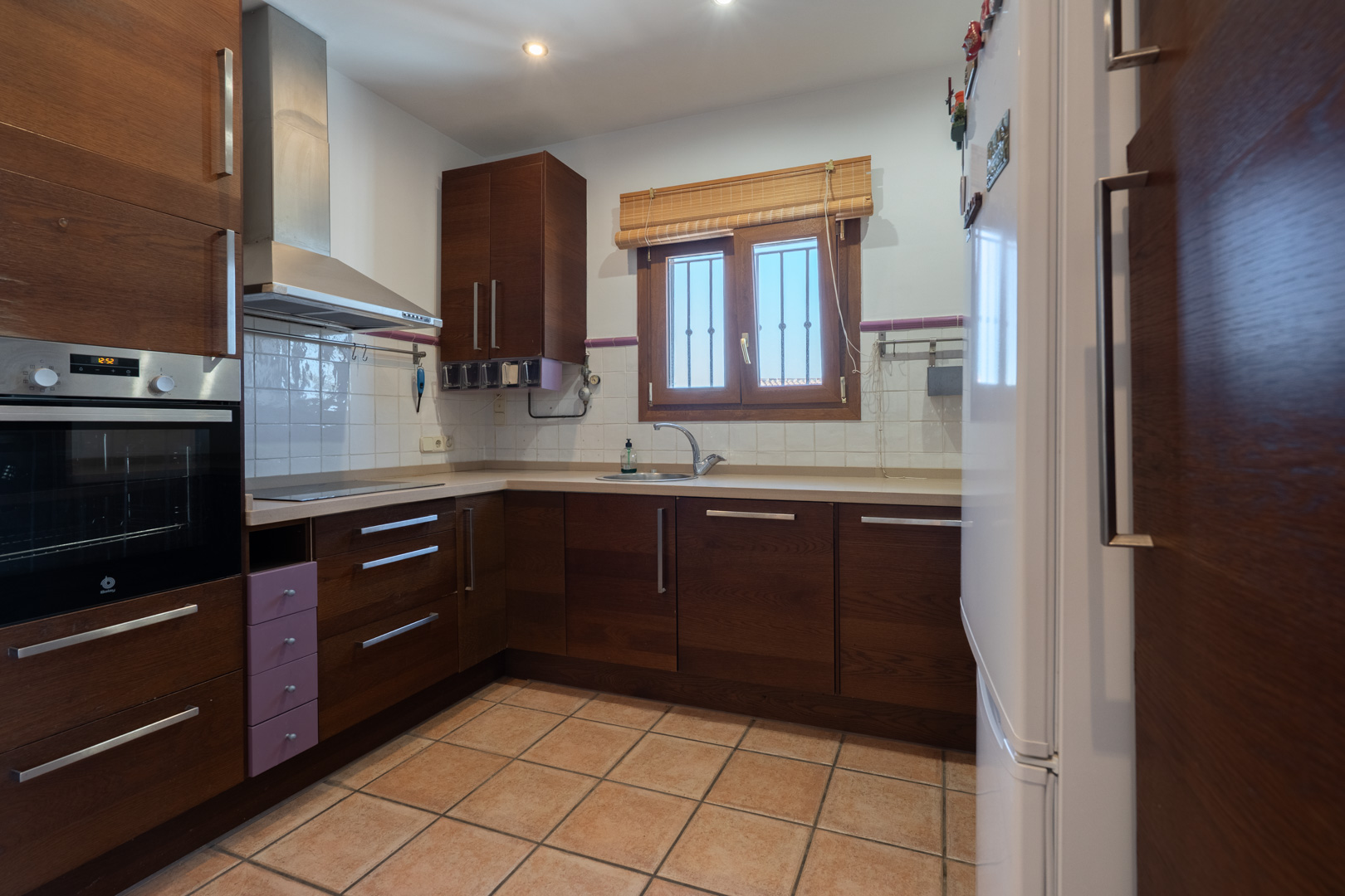 Funktionale Küche mit dunklen Holzschränken, integrierten Geräten und Fliesenboden. Ein Fenster über der Spüle sorgt für natürliches Tageslicht.