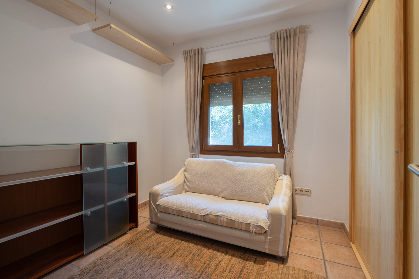 Funktionaler Raum mit Terrakottafliesen, Holzfenster mit Rollläden und Einbauschrank. Ausgestattet mit Wandregalen, Deckenspots und einem kleinen Sofa.
