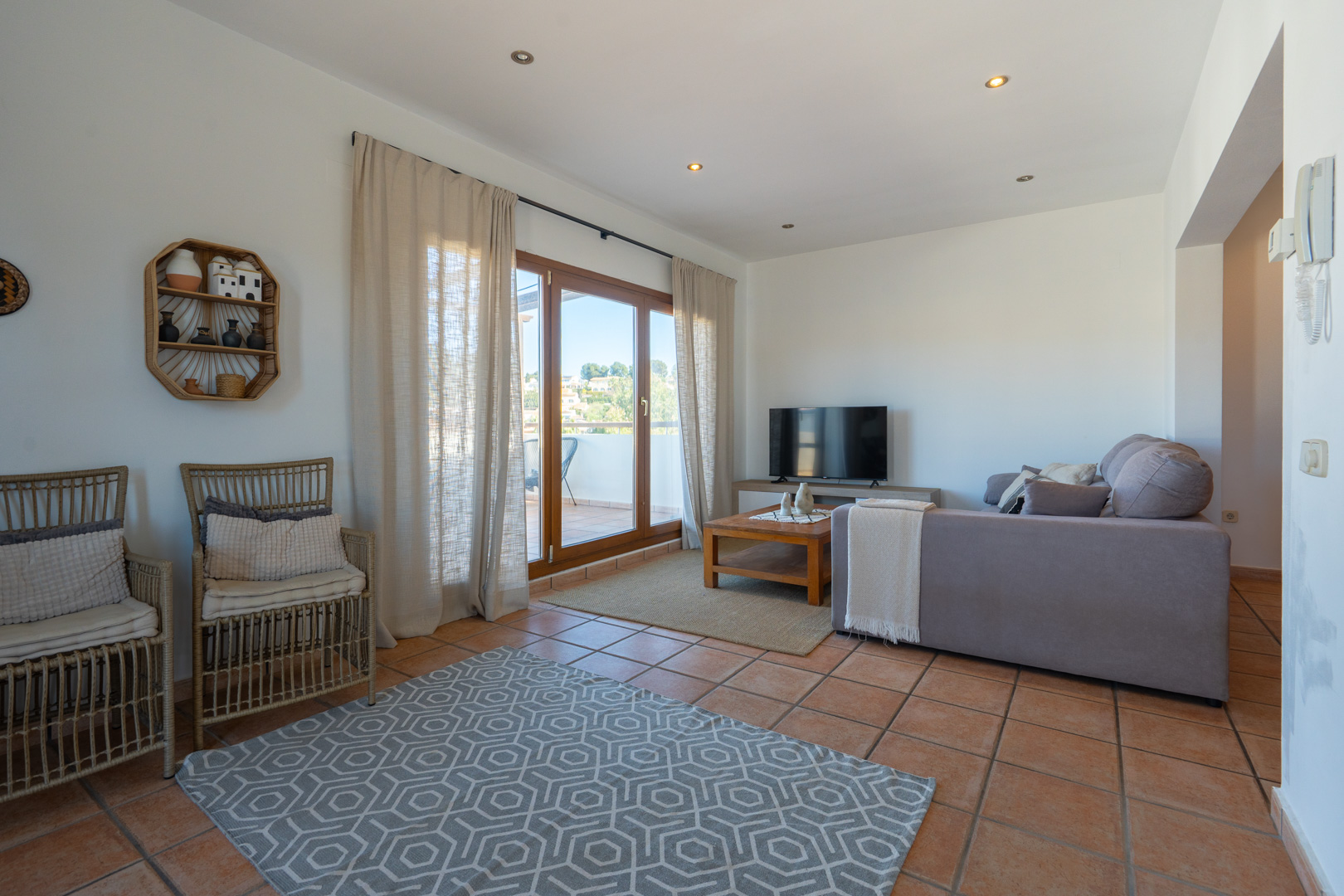 Helles Wohnzimmer mit Terrakottaboden, grauem Ecksofa und direktem Zugang zur Terrasse durch große Glasschiebetüren mit Holzrahmen.
