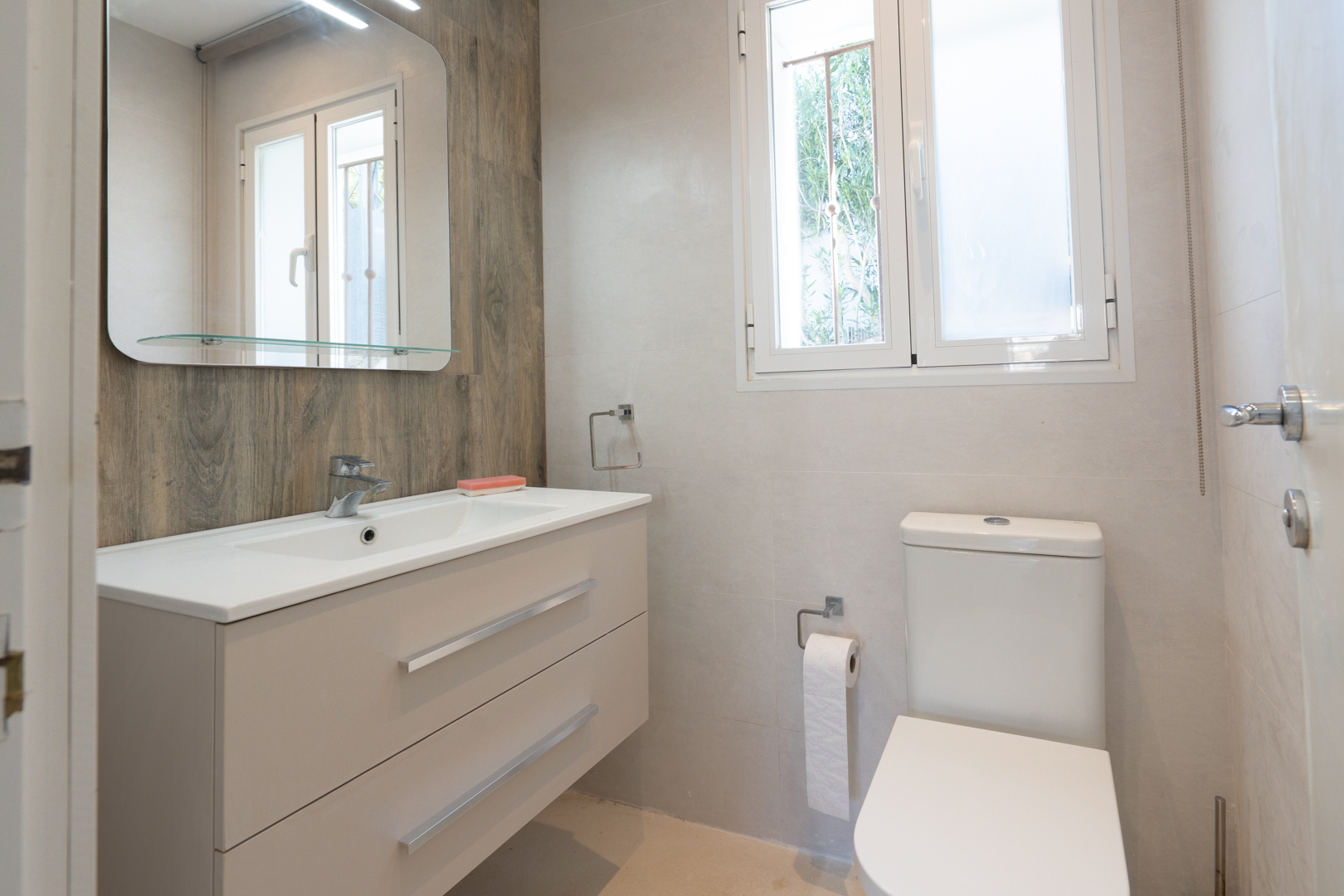 Modernes Badezimmer mit hängendem Waschtisch, Akzentwand in Holzoptik und weißem WC. Ein zweiflügeliges Fenster sorgt für natürliches Licht und Belüftung.