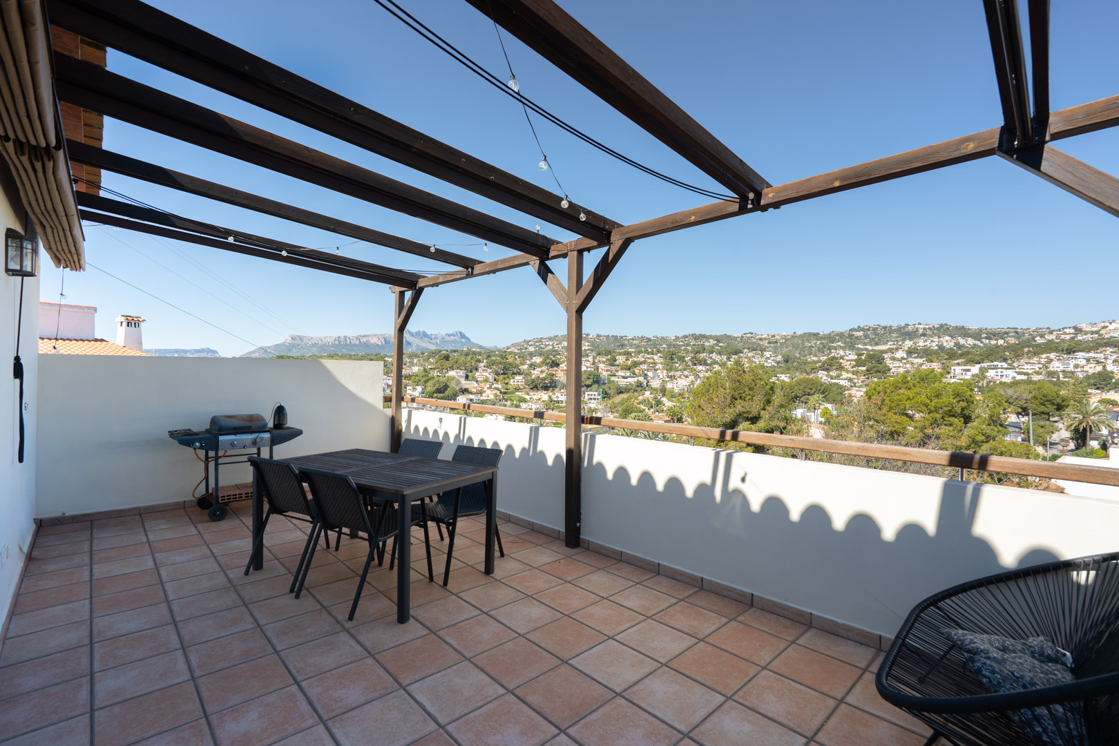 Obere Terrasse mit Terrakotta-Fliesen, Holzpergola und Essbereich. Sie bietet einen weiten Blick auf die Sierra de Bernia und die umliegende Wohngegend.