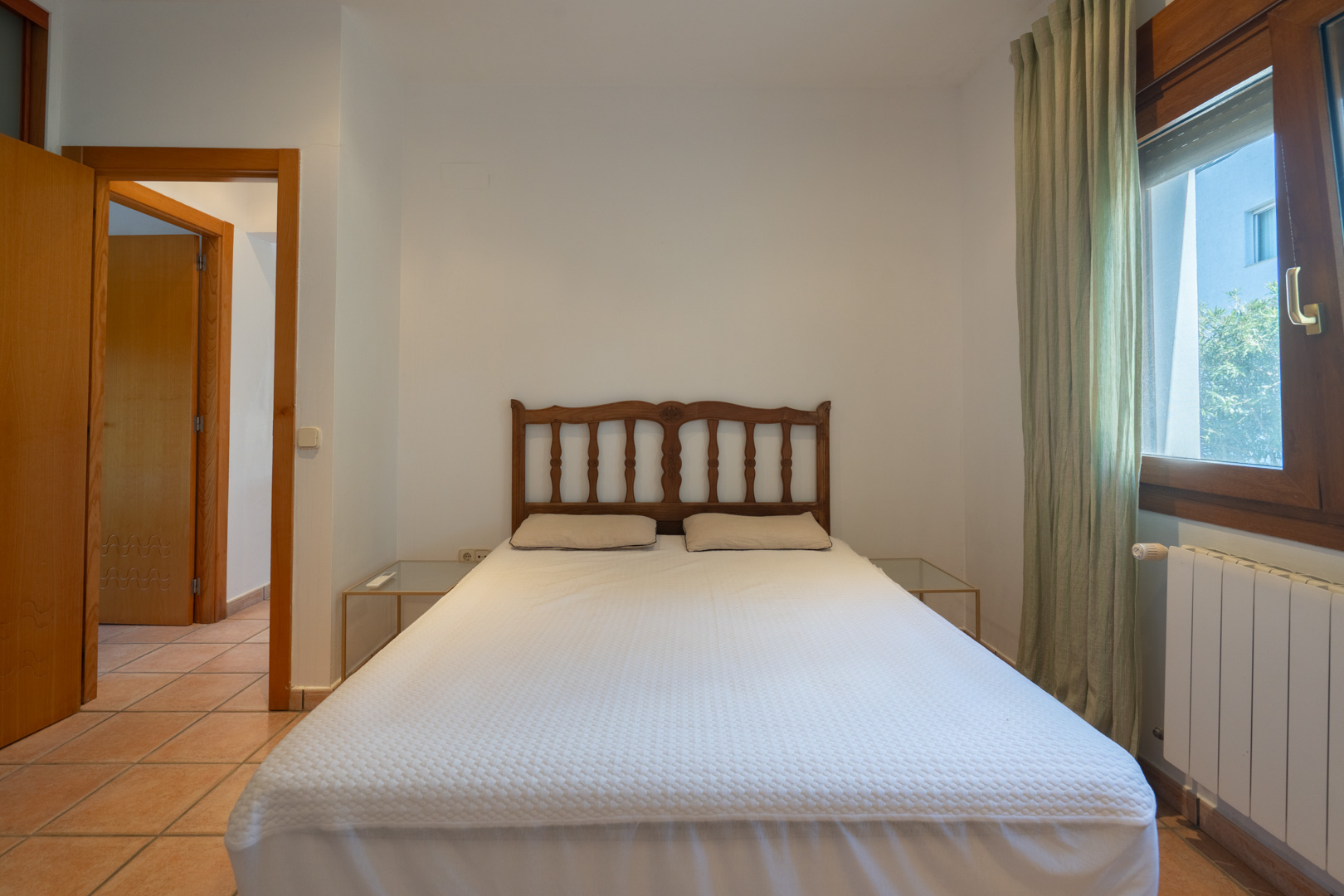 Schlafzimmer mit Doppelbett, geschnitztem Holzkopfteil und Glastischen. Der Raum ist mit Fliesenboden, einem Heizkörper und grünen Vorhängen ausgestattet.
