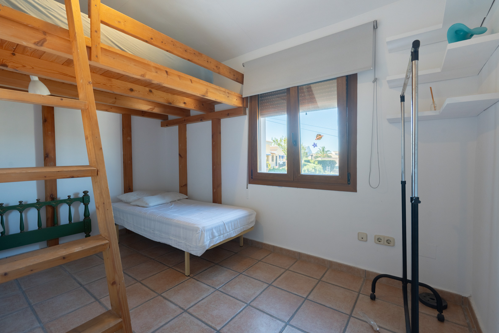 Schlafzimmer mit Hochbettkonstruktion aus Holz, Terrakotta-Fliesenboden und Holzfensterrahmen. Mit weißen Wandregalen und natürlichem Lichteinfall.