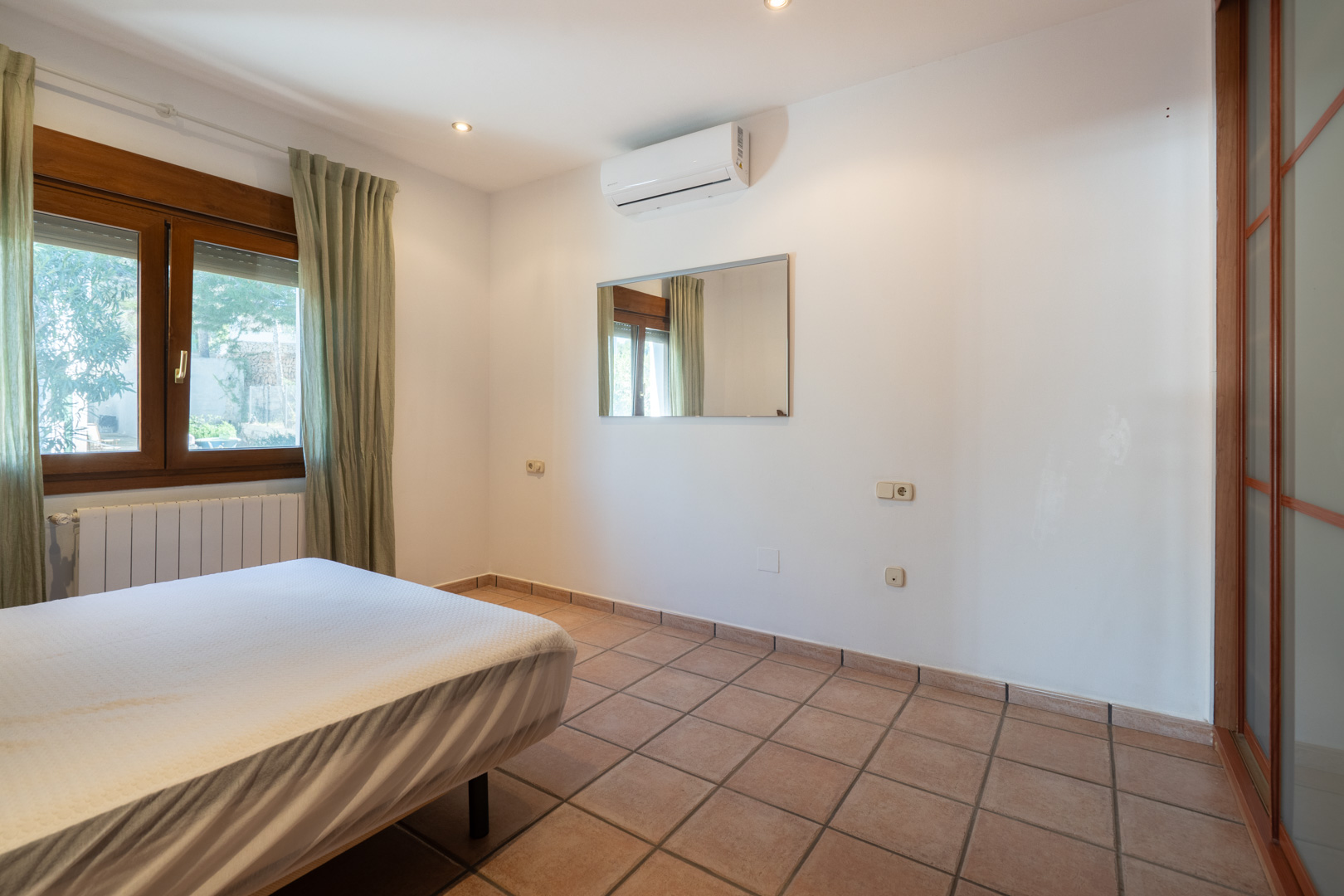 Schlafzimmer mit Terrakotta-Fliesenboden, Split-Klimaanlage und Holzfenster. Der Raum ist mit Deckenspots und einem Schiebetürenschrank ausgestattet.