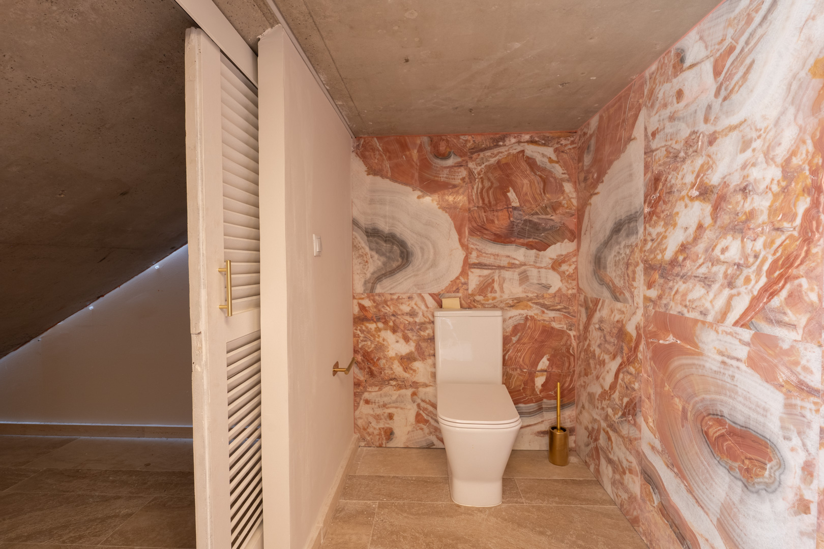 Toilettenraum mit rosa gemaserten Marmorwänden, modernem WC und Sichtbetondecke. Ausgestattet mit einer weißen Lamellen-Schiebetür und goldfarbenen Details.