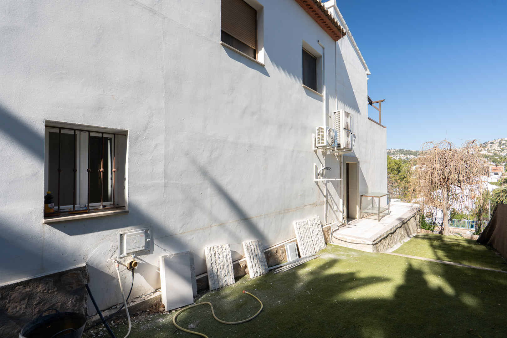 Weiße Seitenfassade mit Natursteinsockel, externen Klimaanlagen und Gartenbereich mit Kunstrasen sowie weitem Blick auf die umliegende Landschaft.