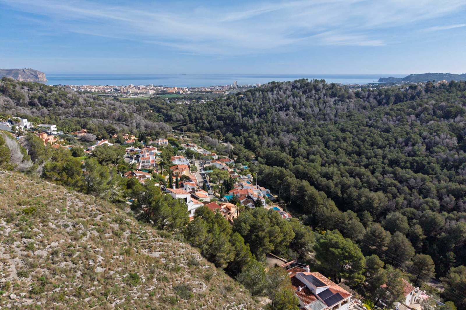 Panorama-Luftaufnahme der erhöhten Lage über bewaldeten Hügeln, mit weitem Blick auf das Mittelmeer und die Silhouette der Küstenstadt Jávea.