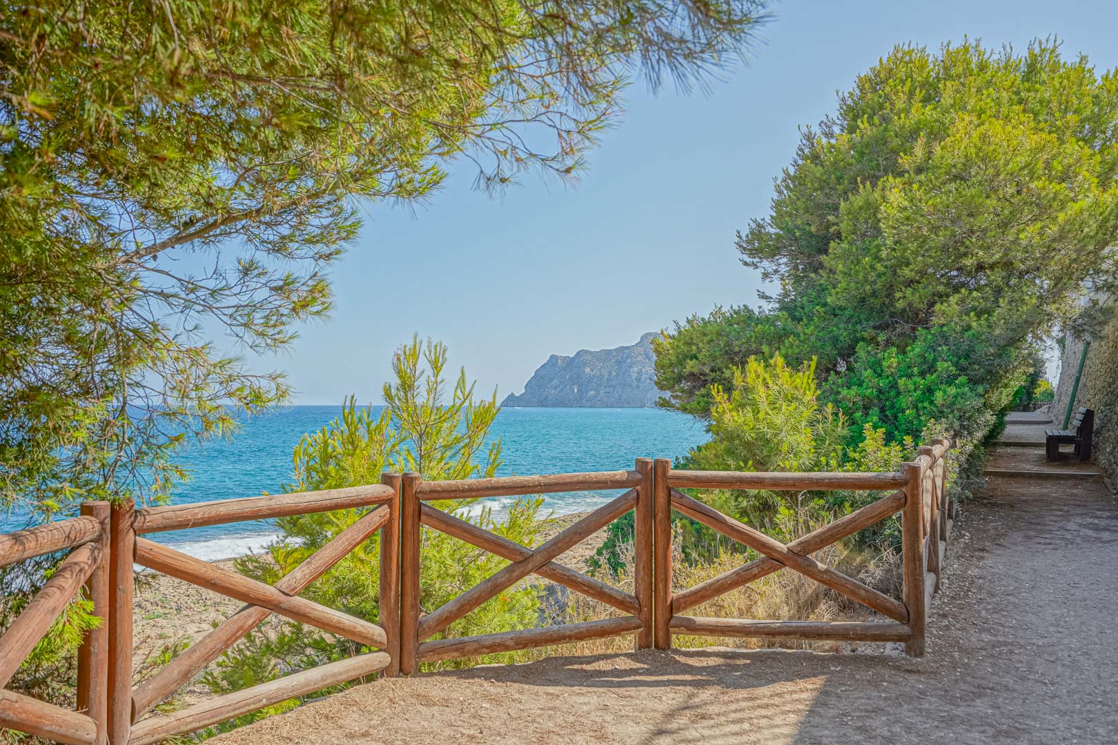 Küstenwanderweg mit rustikalem Holzgeländer und Panoramablick auf das Mittelmeer und den Peñón de Ifach, eingerahmt von Pinien.