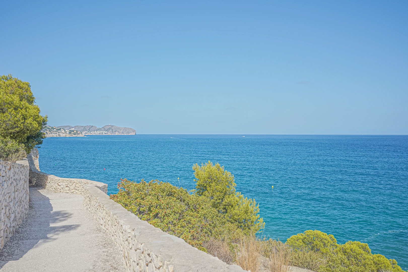Küstenwanderweg mit Trockensteinmauern und Panoramablick auf das Mittelmeer, gesäumt von einheimischen Pinien und mediterraner Flora.