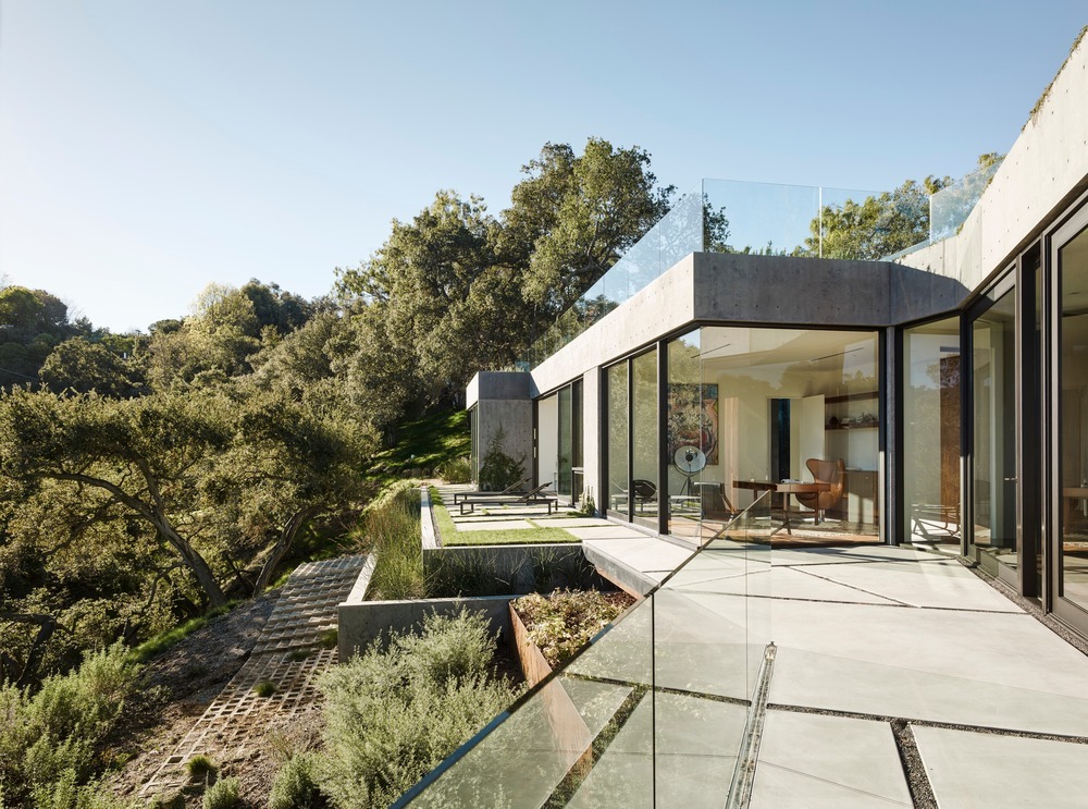 Moderne Betonvilla mit bodentiefen Glasfronten und einer großzügigen Terrasse mit Glasgeländer und geometrischen Steinplatten.