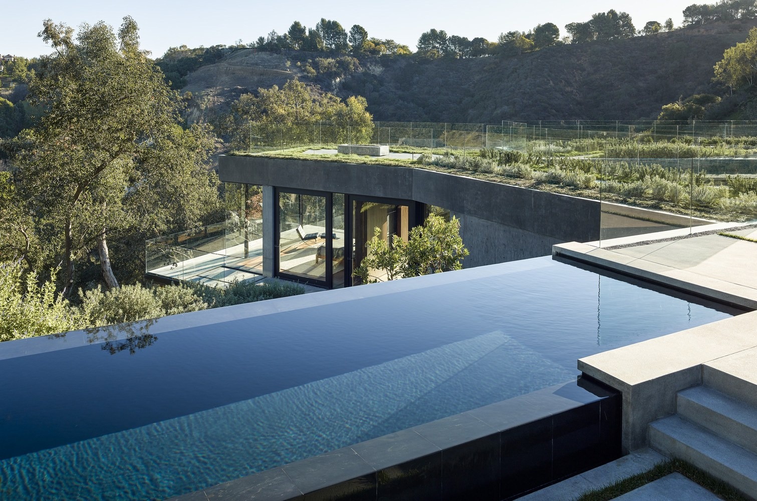 Moderner Infinity-Pool mit dunklen Fliesen, integriert in eine Sichtbeton-Architektur mit Gründach und bodentiefen Glasfronten in Hanglage.