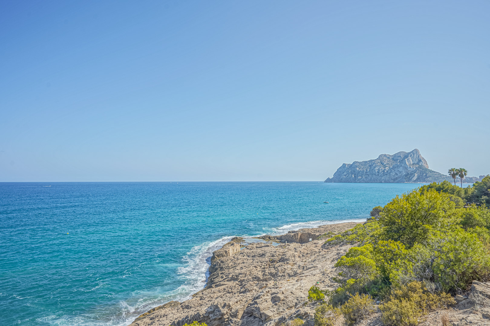 Panoramablick auf die Küste von Benissa mit dem Peñón de Ifach im Hintergrund, kristallklarem türkisfarbenem Wasser und mediterraner Vegetation an den Felsen.