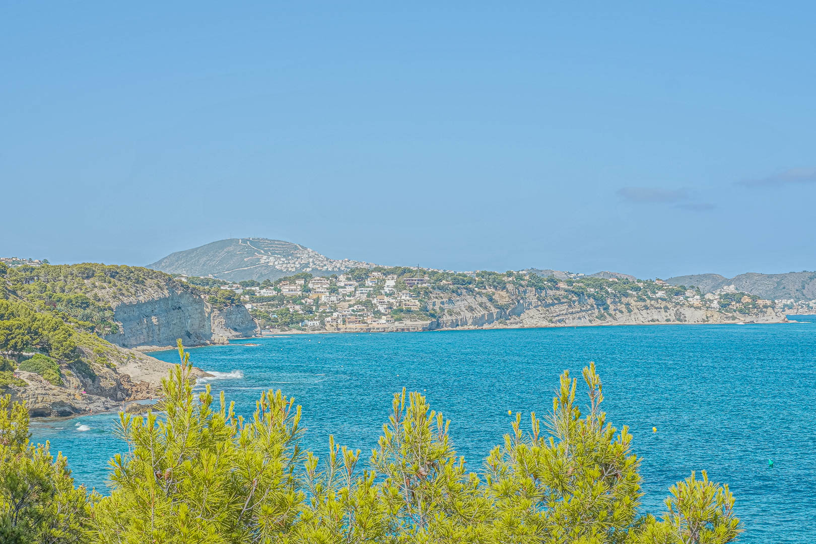 Panoramablick auf die Küste von Benissa mit mediterranen Pinien im Vordergrund und bewohnten Klippen über dem türkisfarbenen Mittelmeer.