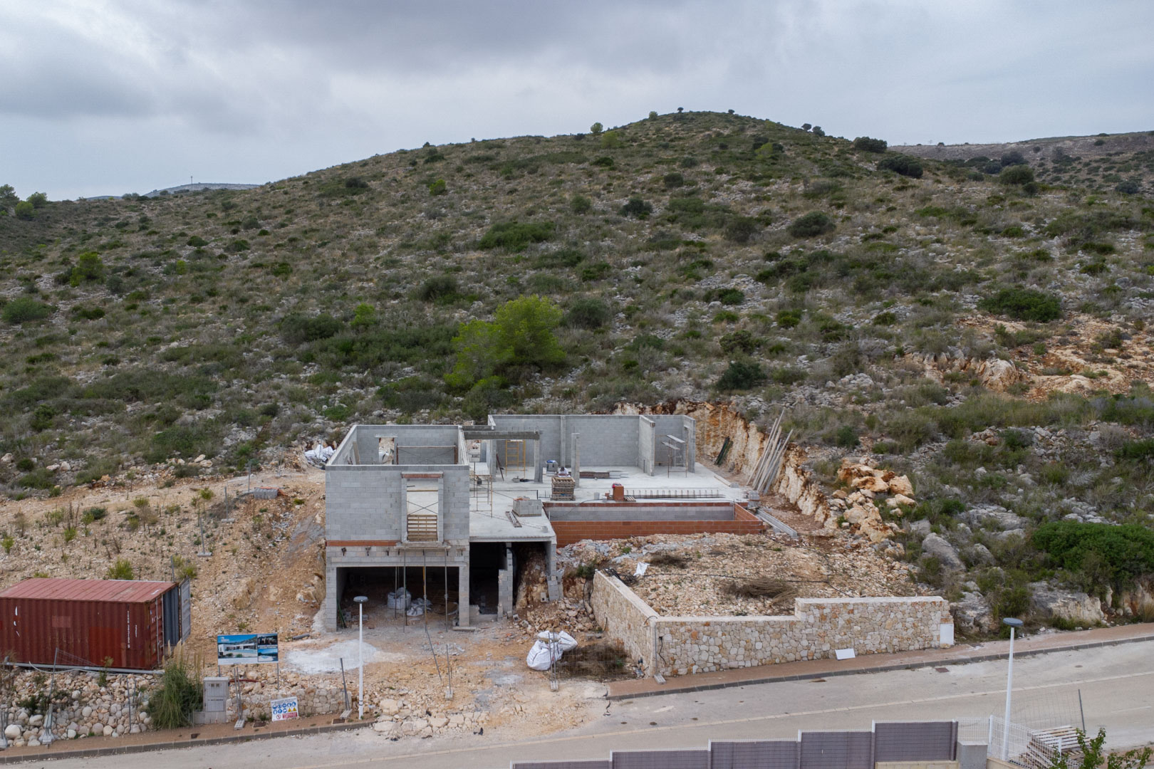 Bauphase einer Villa in Pedreguer mit Betonblockstrukturen, einer Natursteinmauer und der Integration in die felsige Hanglage unter bewölktem Himmel.