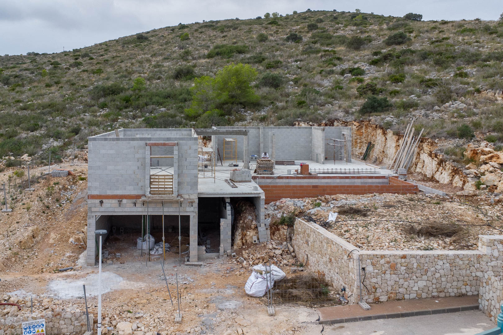 Rohbau einer modernen Villa in Pedreguer mit Betonblock-Struktur, Naturstein-Stützmauern und Hanglage inmitten der mediterranen Landschaft.