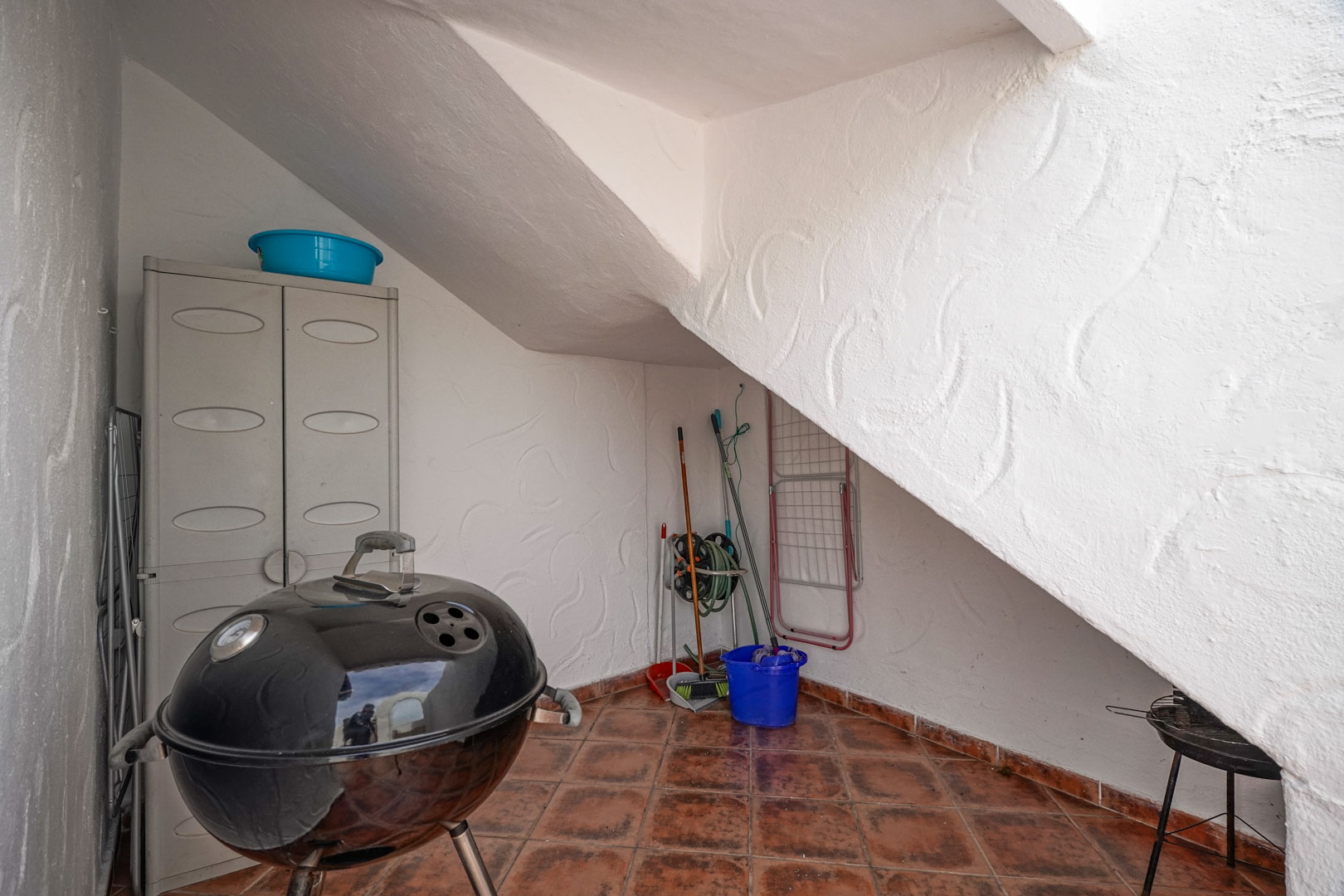 Außenabstellbereich unter der Treppe mit Terrakottafliesen, Kunststoffschrank und Stellfläche für einen Kugelgrill sowie Reinigungsgeräte.