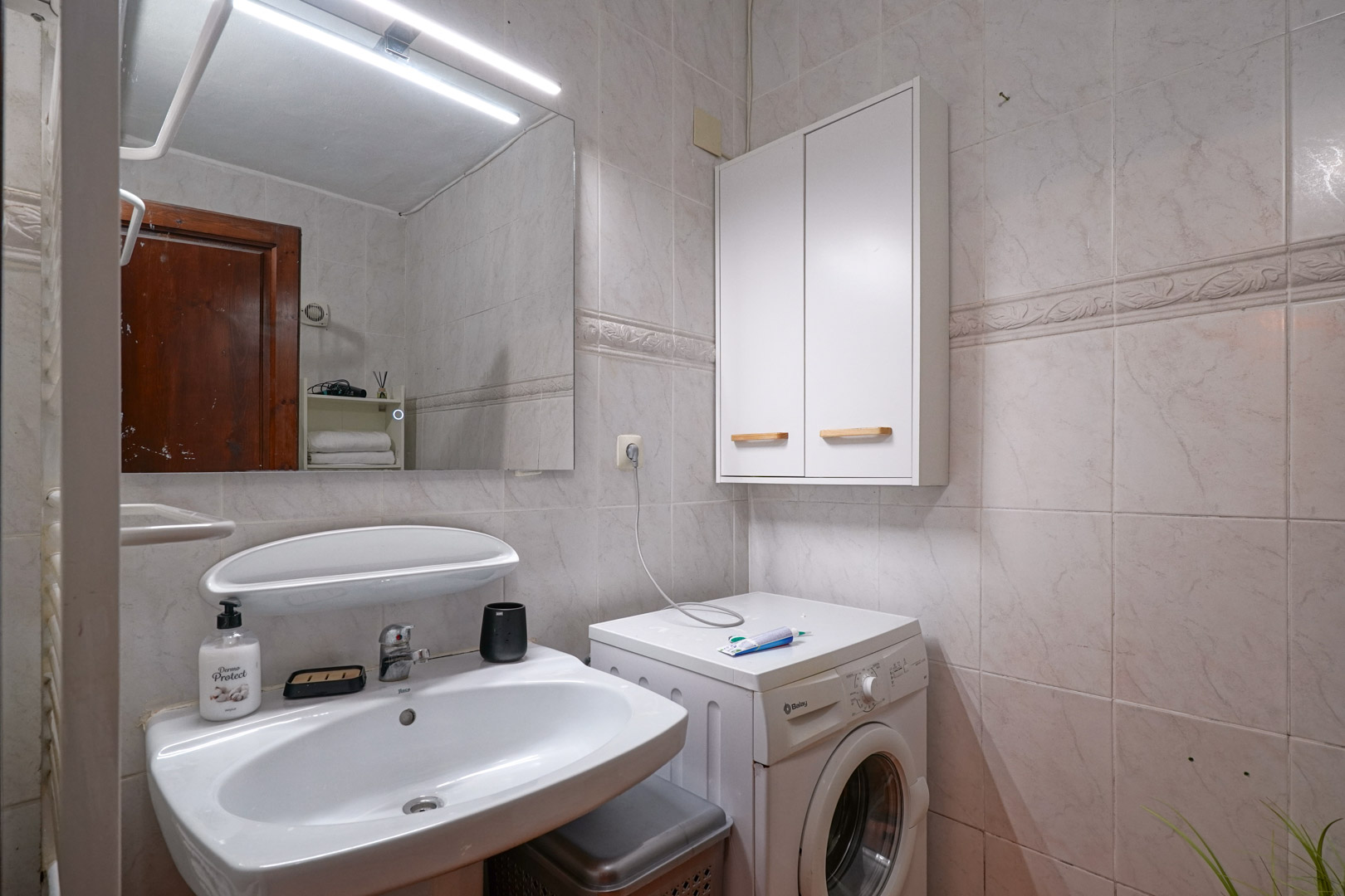 Funktionales Badezimmer mit Keramikwaschbecken, LED-Spiegel und integriertem Wäschebereich mit Waschmaschine und Hängeschrank auf hell gefliesten Wänden.