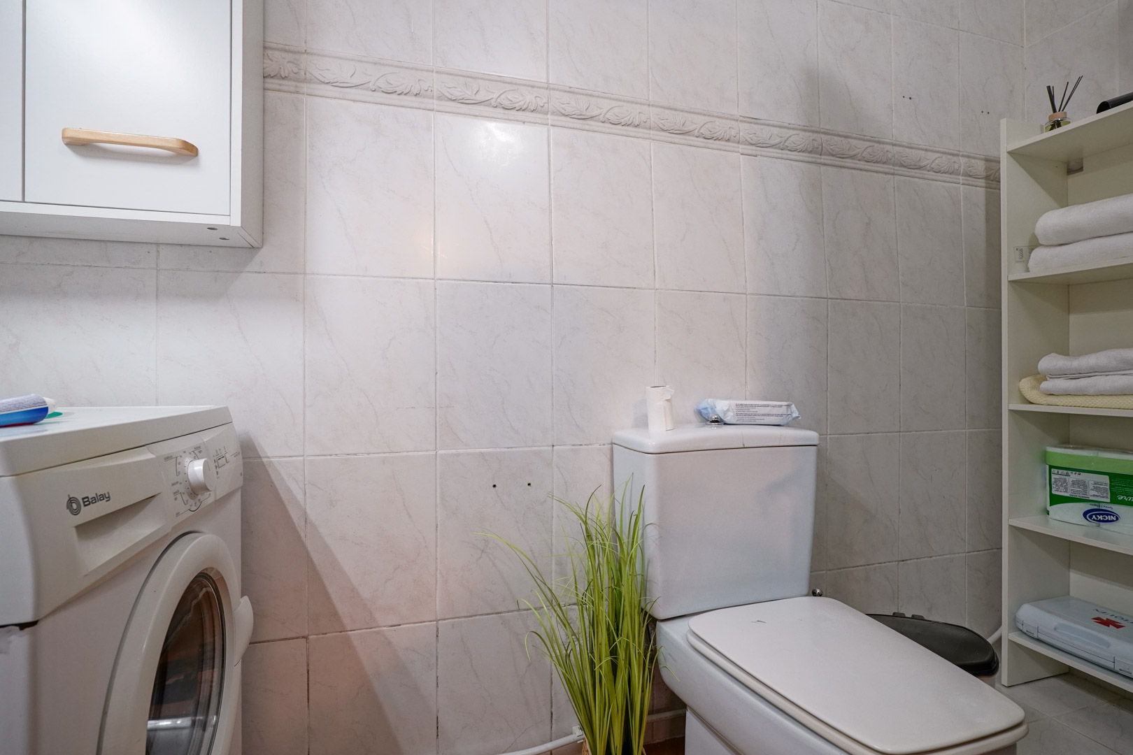 Funktionales Badezimmer mit Wäschebereich, ausgestattet mit einer Balay-Waschmaschine, Lagerregalen und gefliesten Wänden mit Zierborte.
