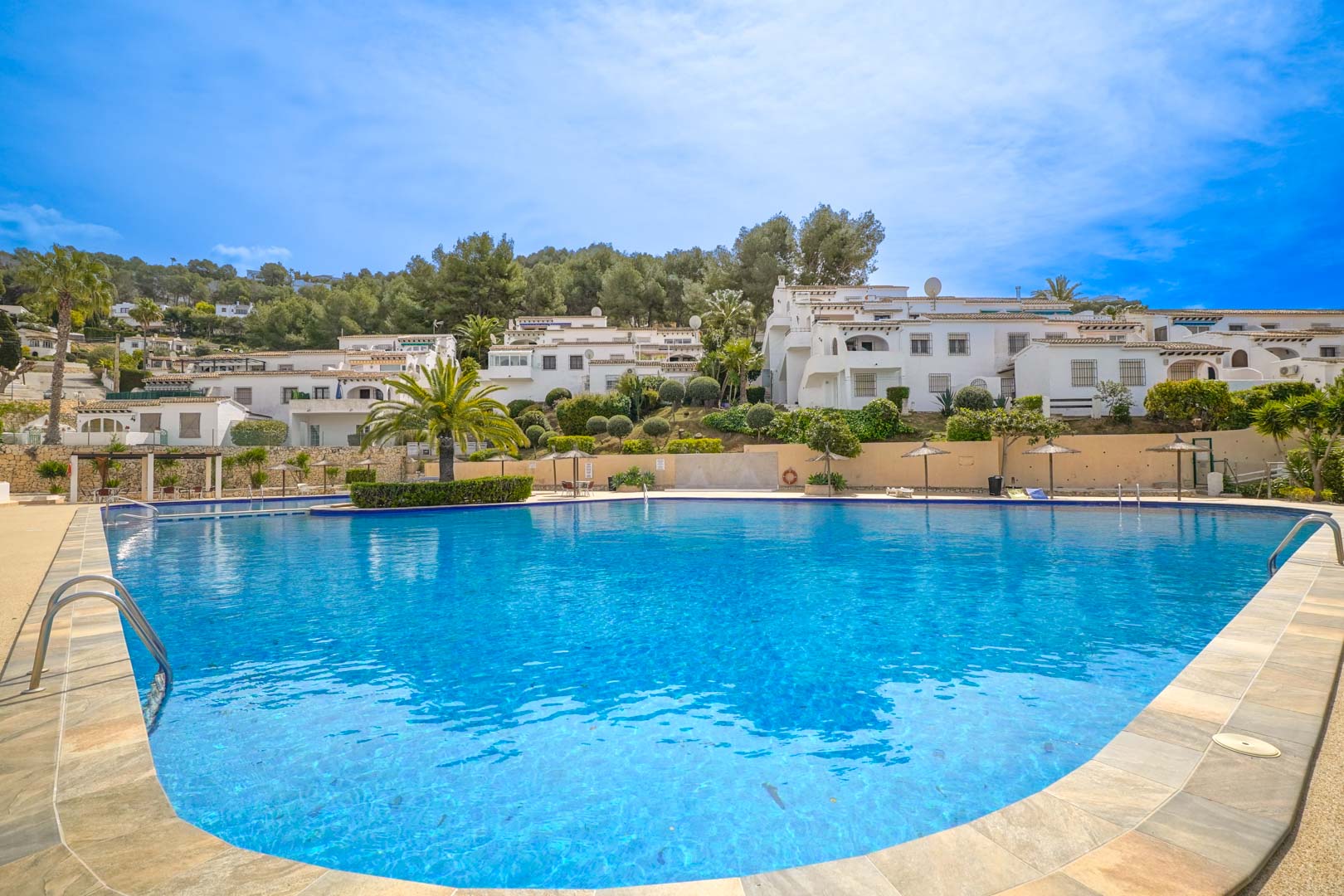 Großer Gemeinschaftspool mit geschwungenen Formen, umgeben von einer Sonnenterrasse mit Strohschirmen und mediterraner Bepflanzung vor der Kulisse weißer Wohngebäude.