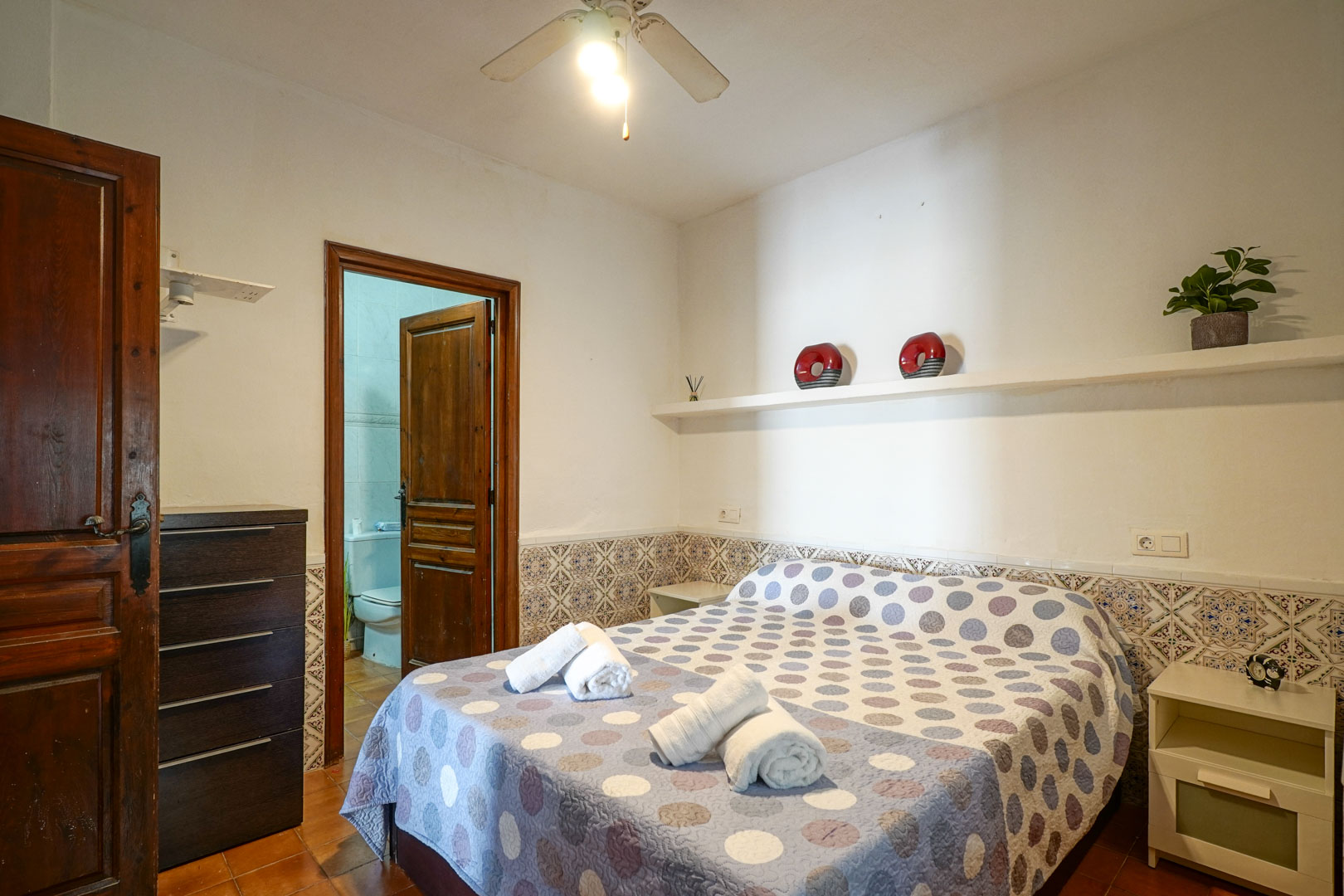 Hauptschlafzimmer mit dekorativem Fliesensockel, Deckenventilator und direktem Zugang zum angrenzenden Badezimmer durch eine rustikale Holztür.