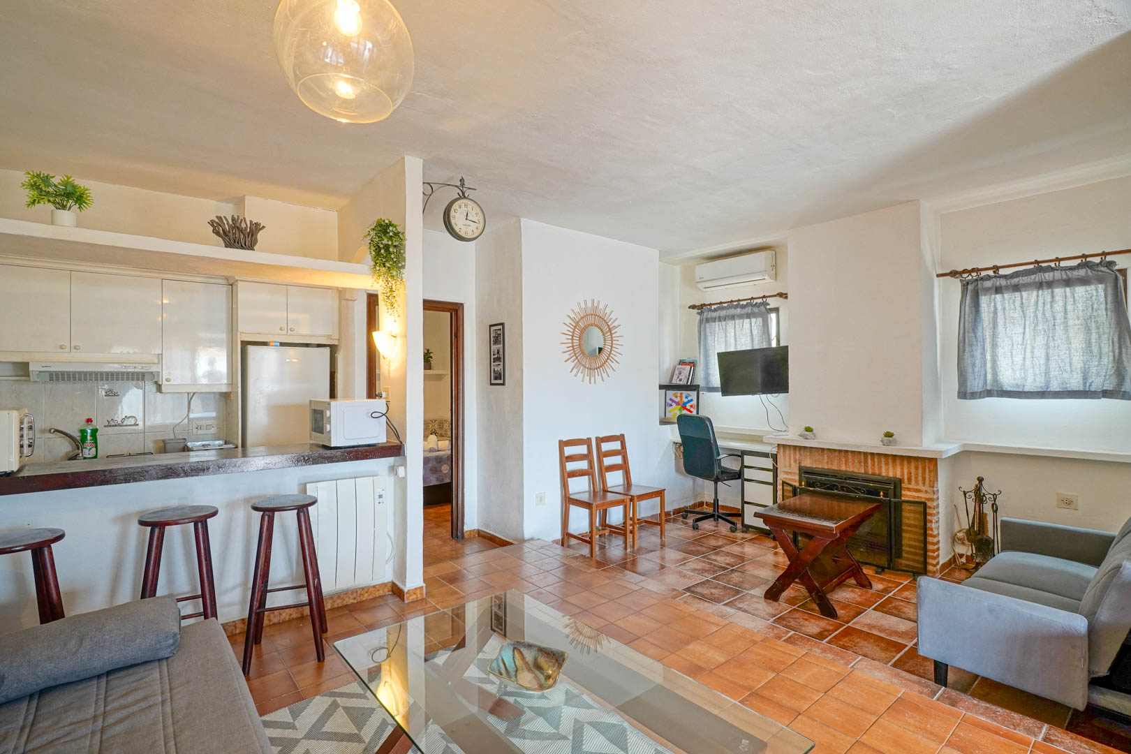 Offener Wohnbereich mit gemauertem Kamin, Terrakottaboden und halb-integrierter Küche mit Frühstückstheke und Holzbarhockern.