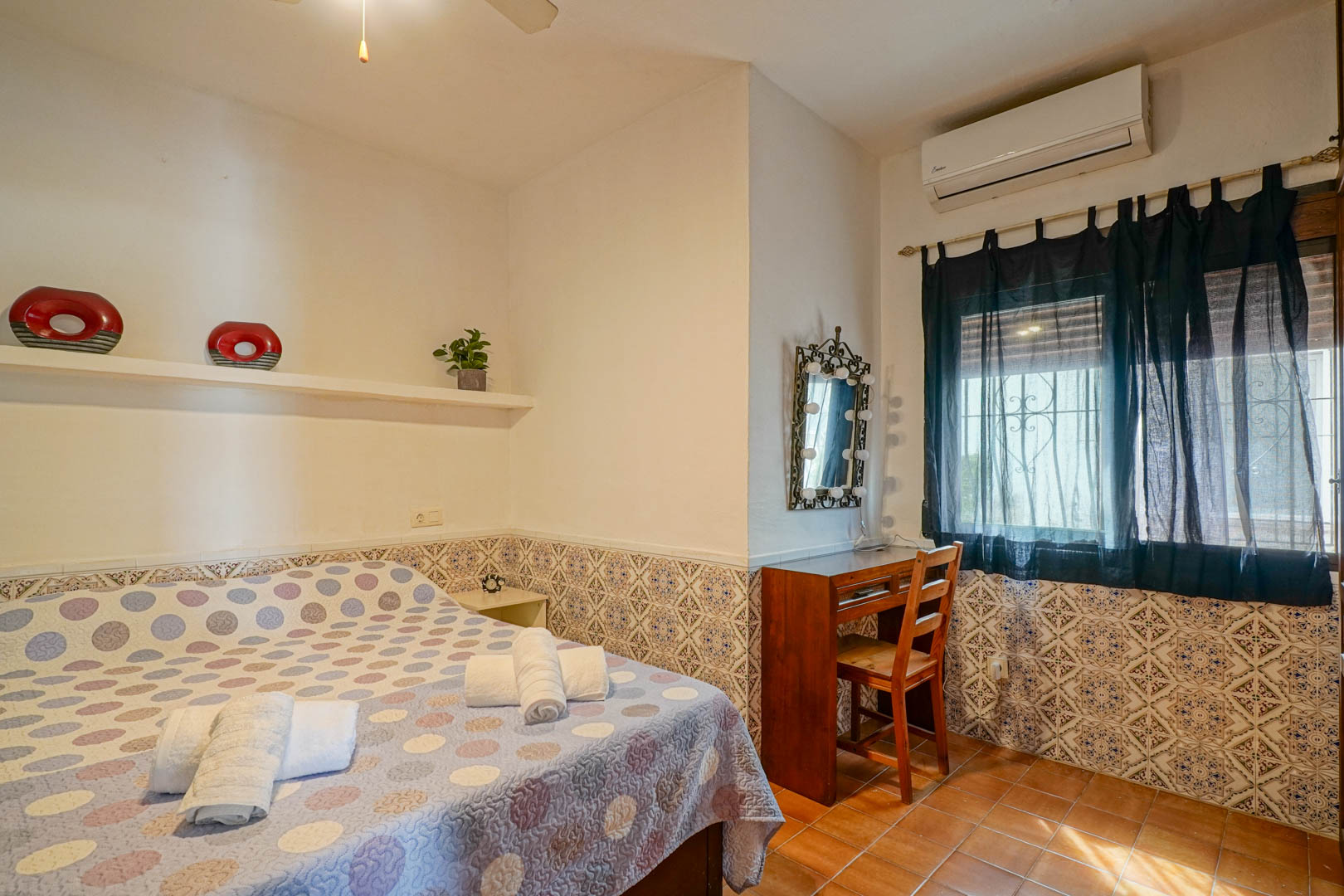 Schlafzimmer mit dekorativer Fliesenumrandung, Holzschreibtisch und beleuchtetem Spiegel. Ausgestattet mit Klimaanlage und rustikalen Bodenfliesen.