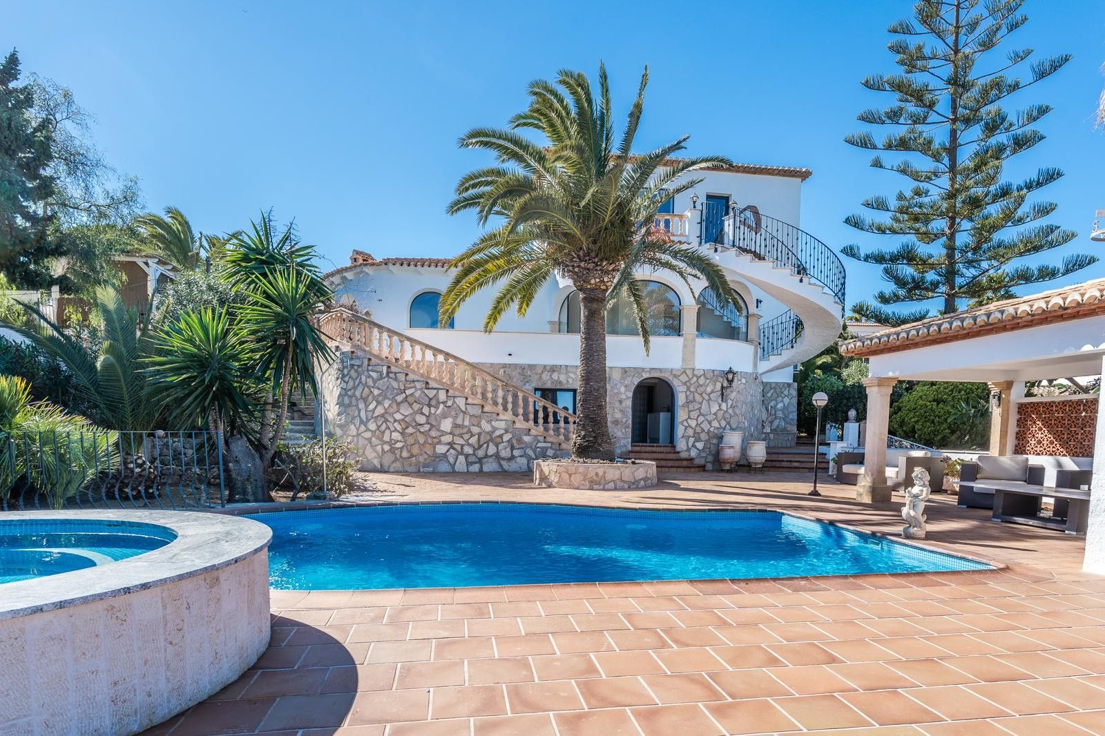 Außenansicht der Villa mit Pool, rundem Whirlpool und Natursteinfassade. Gezeigt werden eine Wendeltreppe und ein mediterraner Garten mit Palmen und Terrassen.