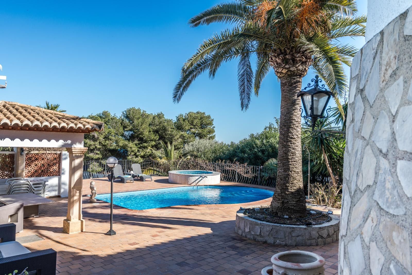 Außenbereich mit geschwungenem Pool, rundem Whirlpool und überdachter Veranda, eingebettet in einen mediterranen Garten mit Palmen und Terrakottaboden.
