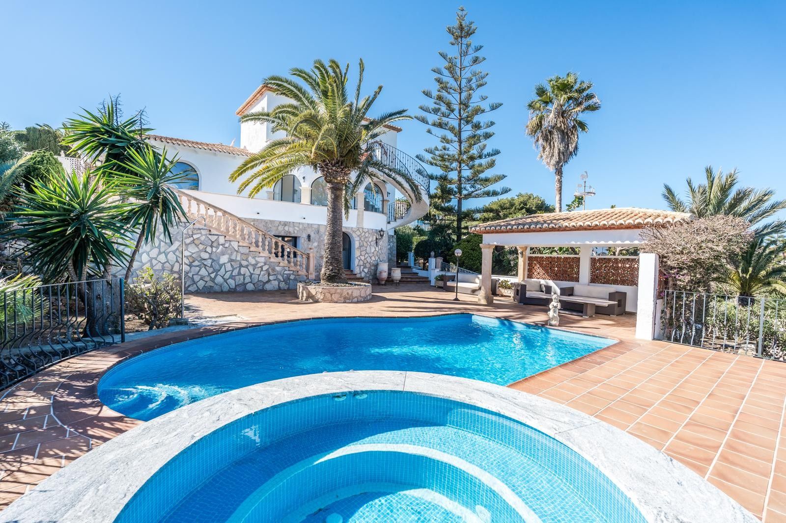 Außenbereich mit renoviertem Pool, rundem Whirlpool und überdachter Lounge. Die weiße Fassade mit Natursteinsockel wird von Palmen und mediterraner Bepflanzung ergänzt.
