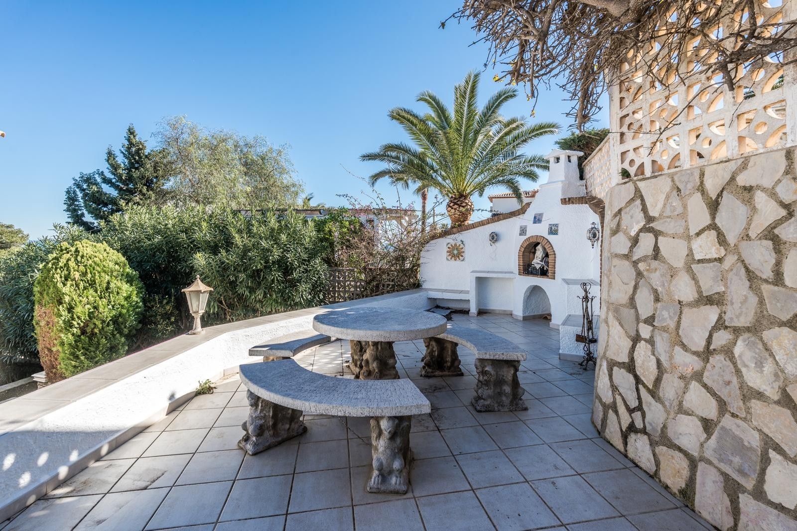 Außenunterhaltungsterrasse mit einem Esstisch-Set aus Naturstein, integriertem gemauertem Grill und Steinmauern in einer mediterranen Gartenanlage.