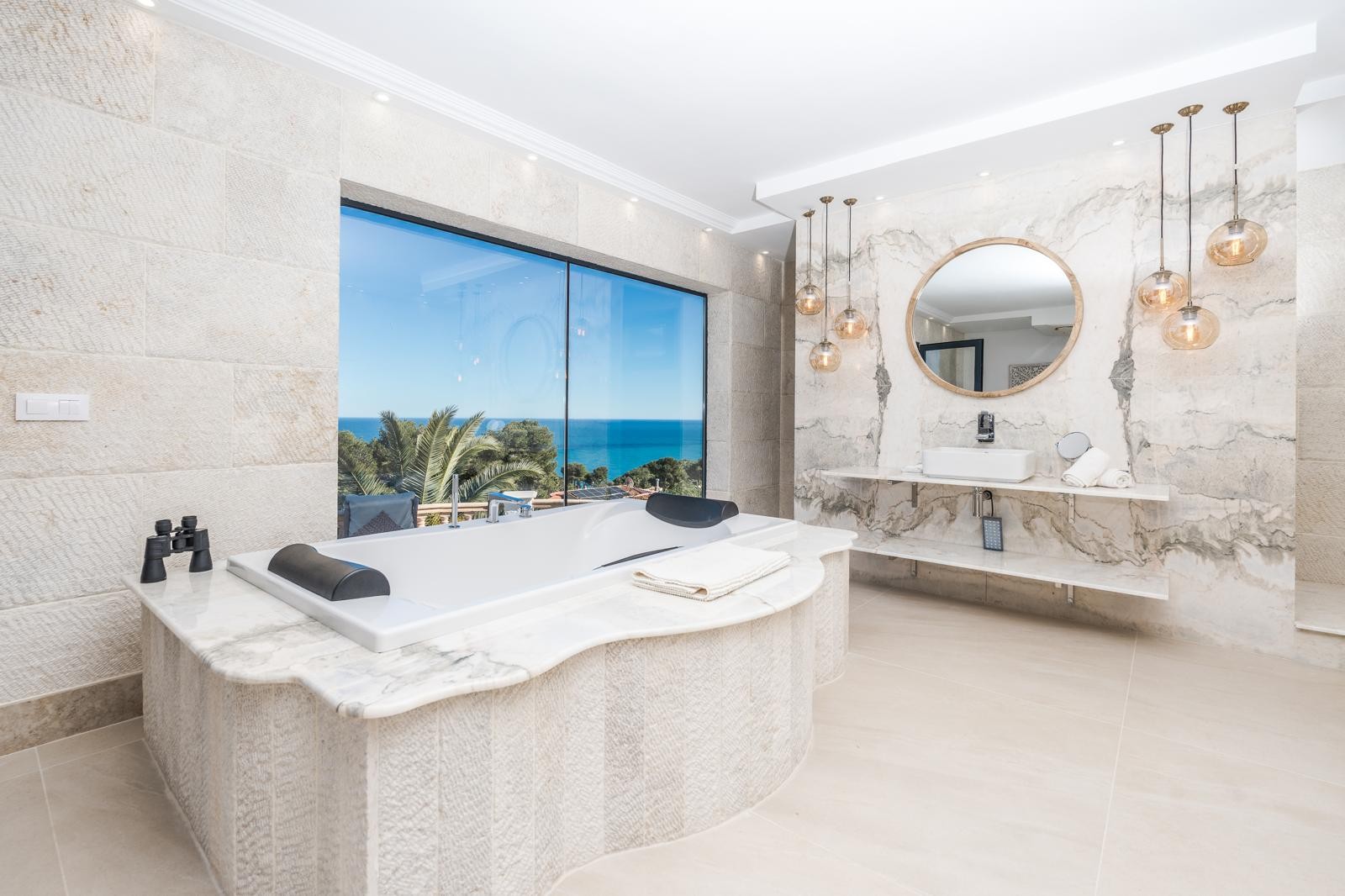 Badezimmer der Master-Suite mit Badewanne vor dem Panoramafenster mit Meerblick, Wänden aus gemasertem Marmor und eleganten Pendelleuchten.