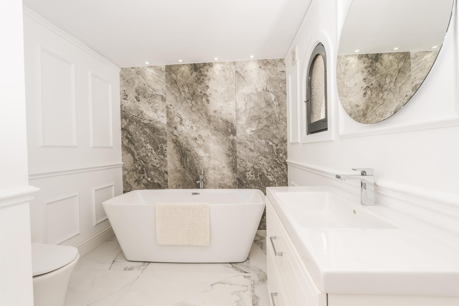 Badezimmer der Master-Suite mit freistehender Badewanne, Marmor-Akzentwand und Wandleisten. Ausgestattet mit modernem Waschtisch, Rundspiegel und einem mediterranen Rundbogenfenster.