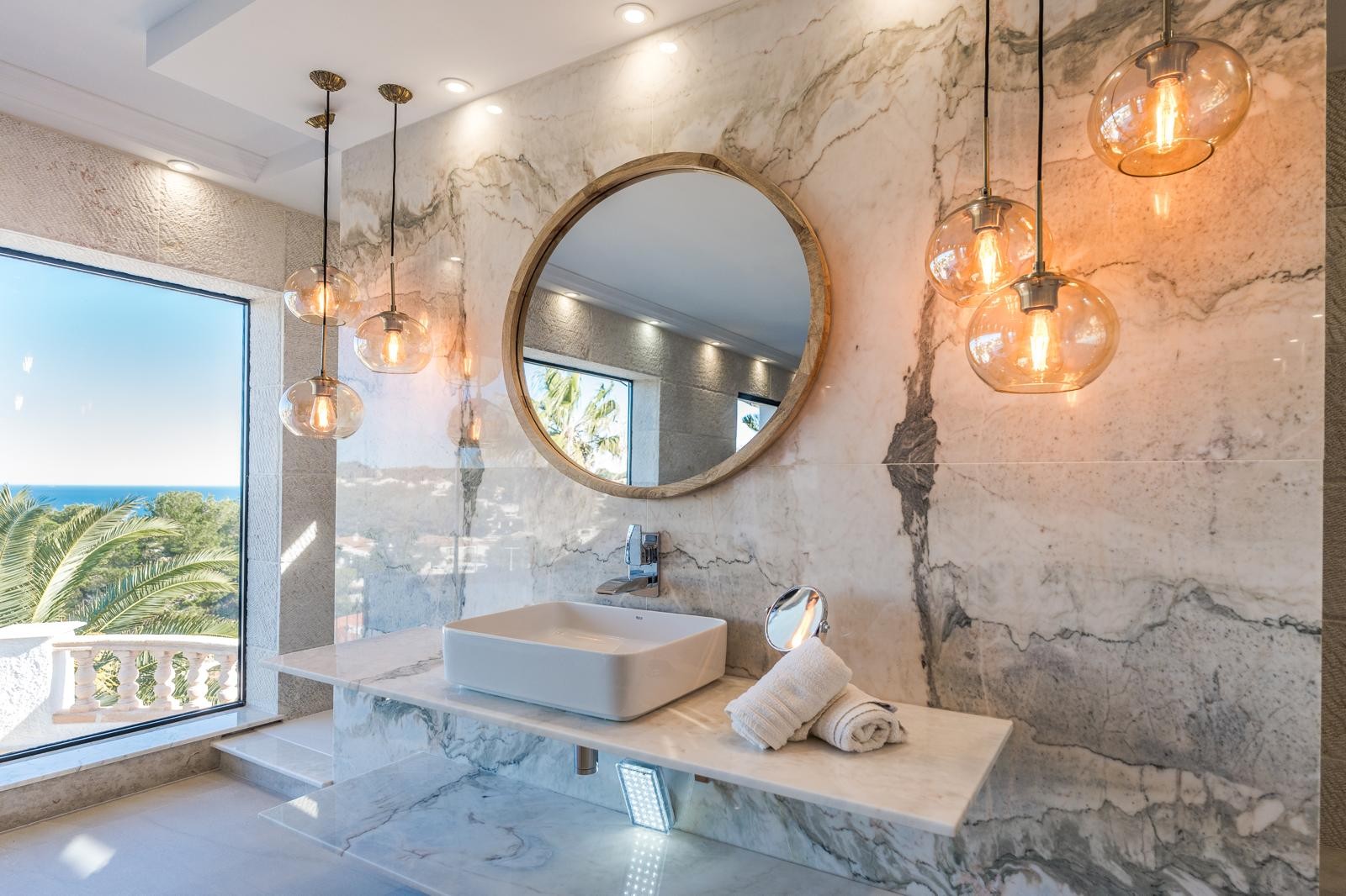 Badezimmer der Master-Suite mit marmorierten Wänden, Aufsatzwaschbecken, rundem Holzspiegel und bodentiefem Fenster mit weitem Blick auf die Küste von Jávea.